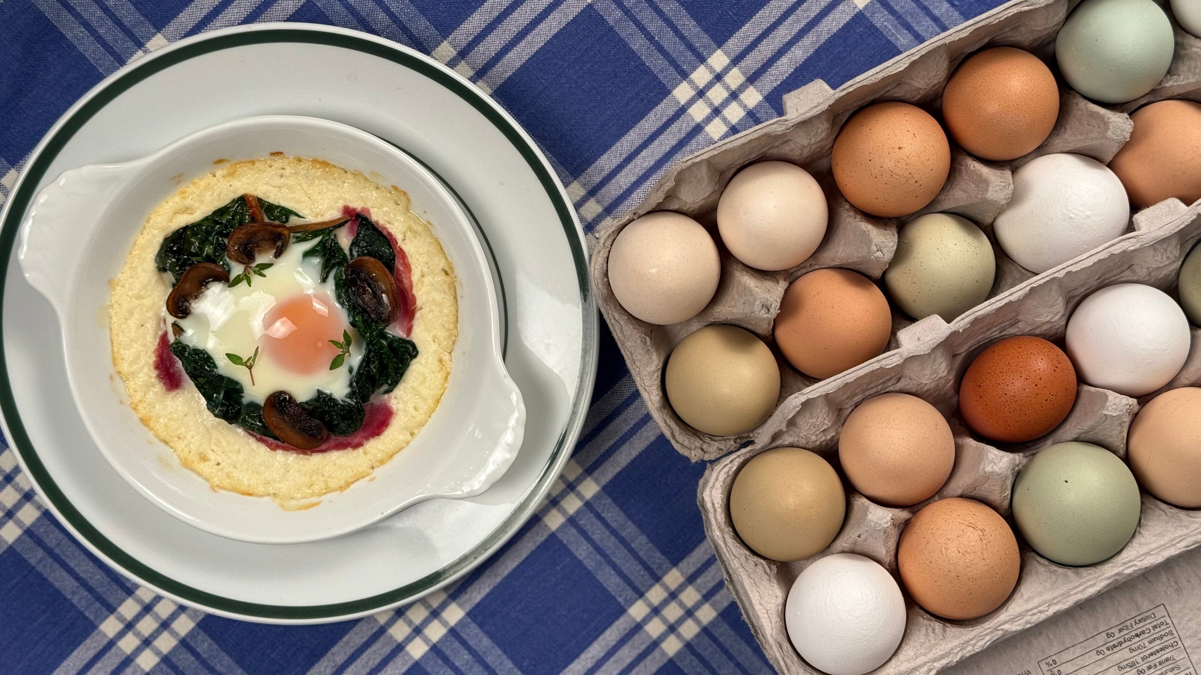 Farmhouse egg dish in a French porcelain gratin, framed by open cartons of farm eggs in varied sizes and shell textures, arranged on a blue plaid cloth.