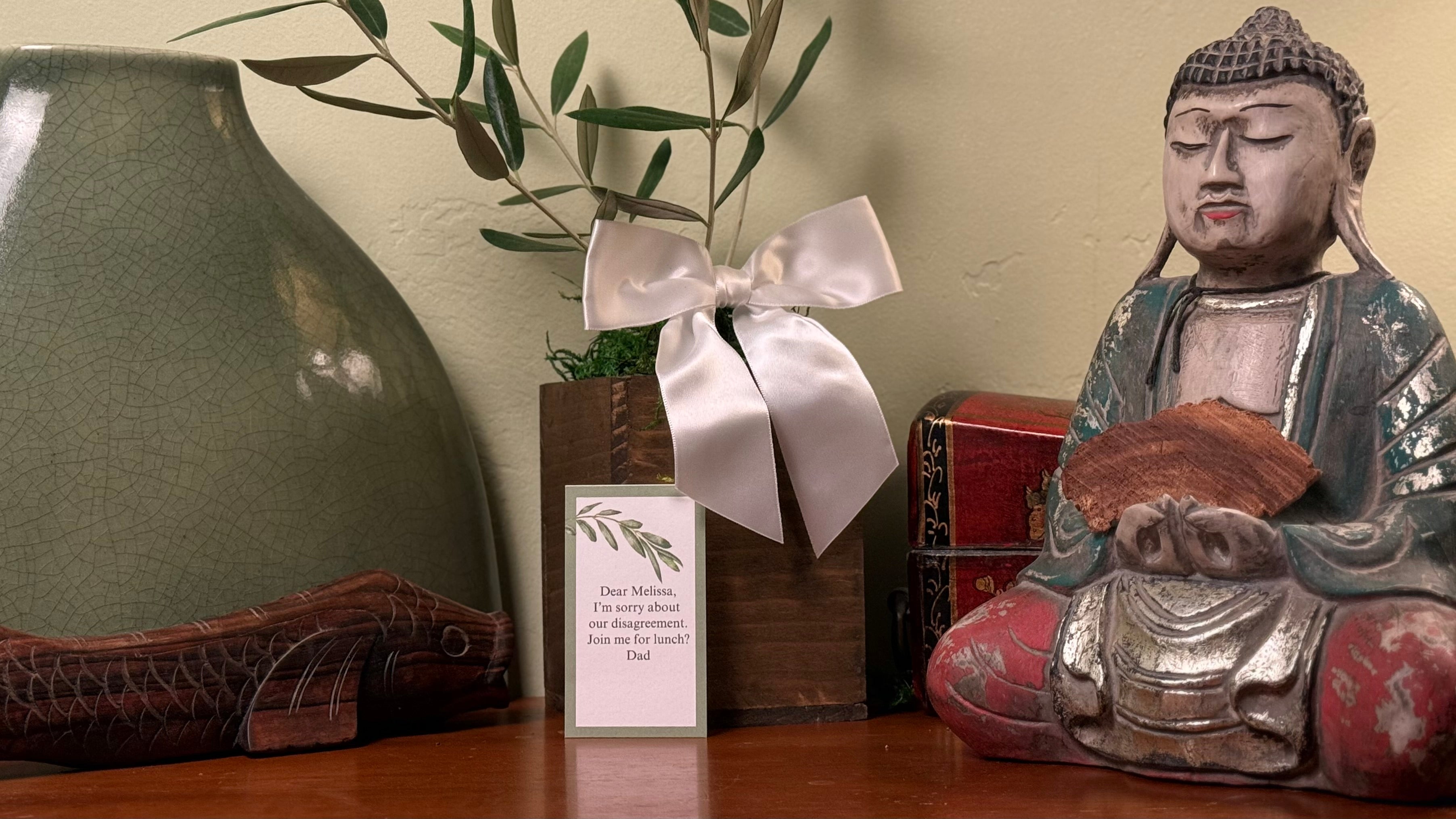 The an olive tree gift in a rustic wooden cachepot with a white bow is on a table next to a small Buddha.