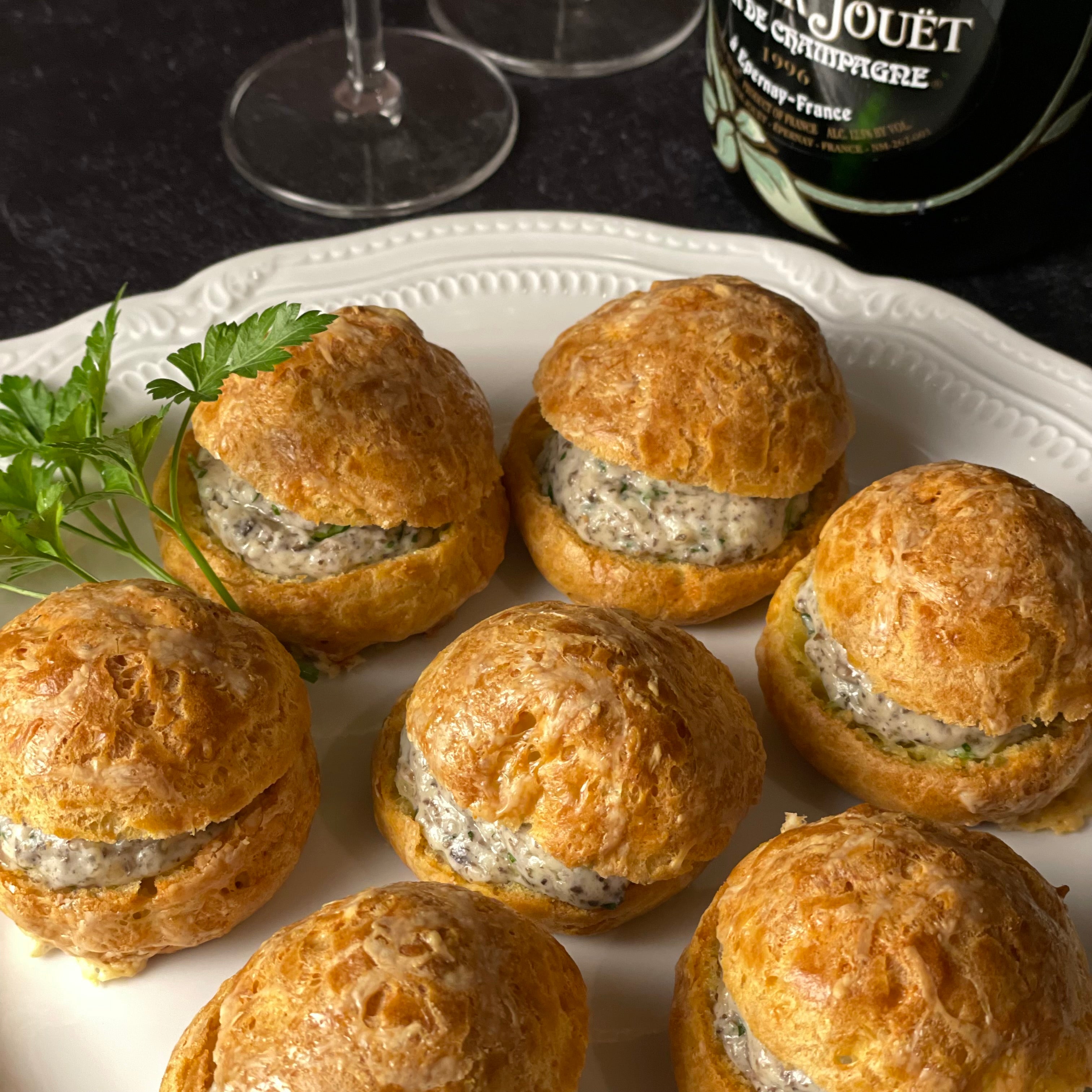 A delicate round Limoges Platter is filled with 7 Gougères with Mornay Duxelles filling. The bottom of a bottle of Perrier-Jouët Belle Epoque is barely seen on the edge of the image.