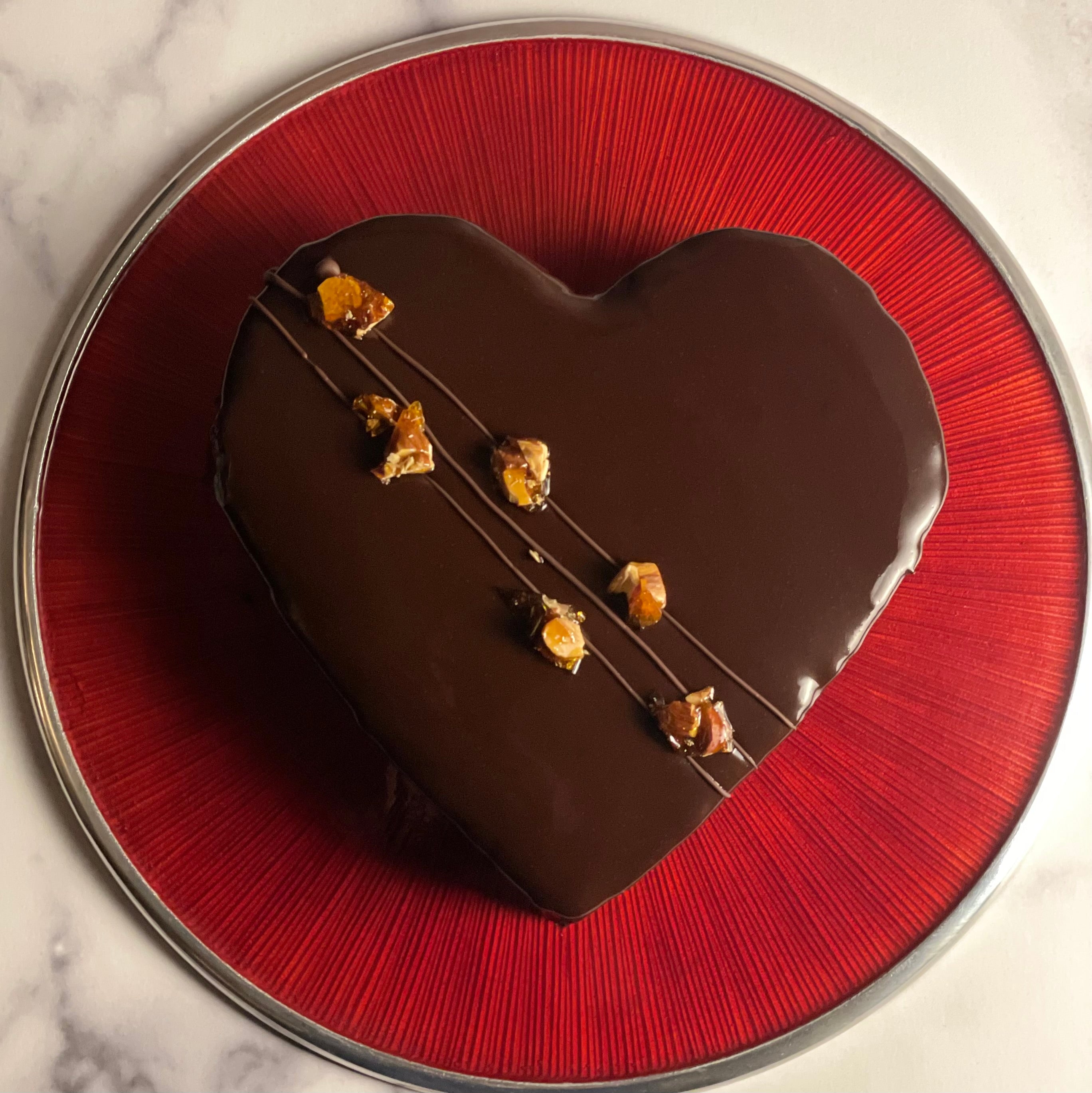 This is an overhead image of a heart shaped chocolate cake with some diagonal chocolate stripes running left to right and 8 little chunks of almond praline placed upon them.