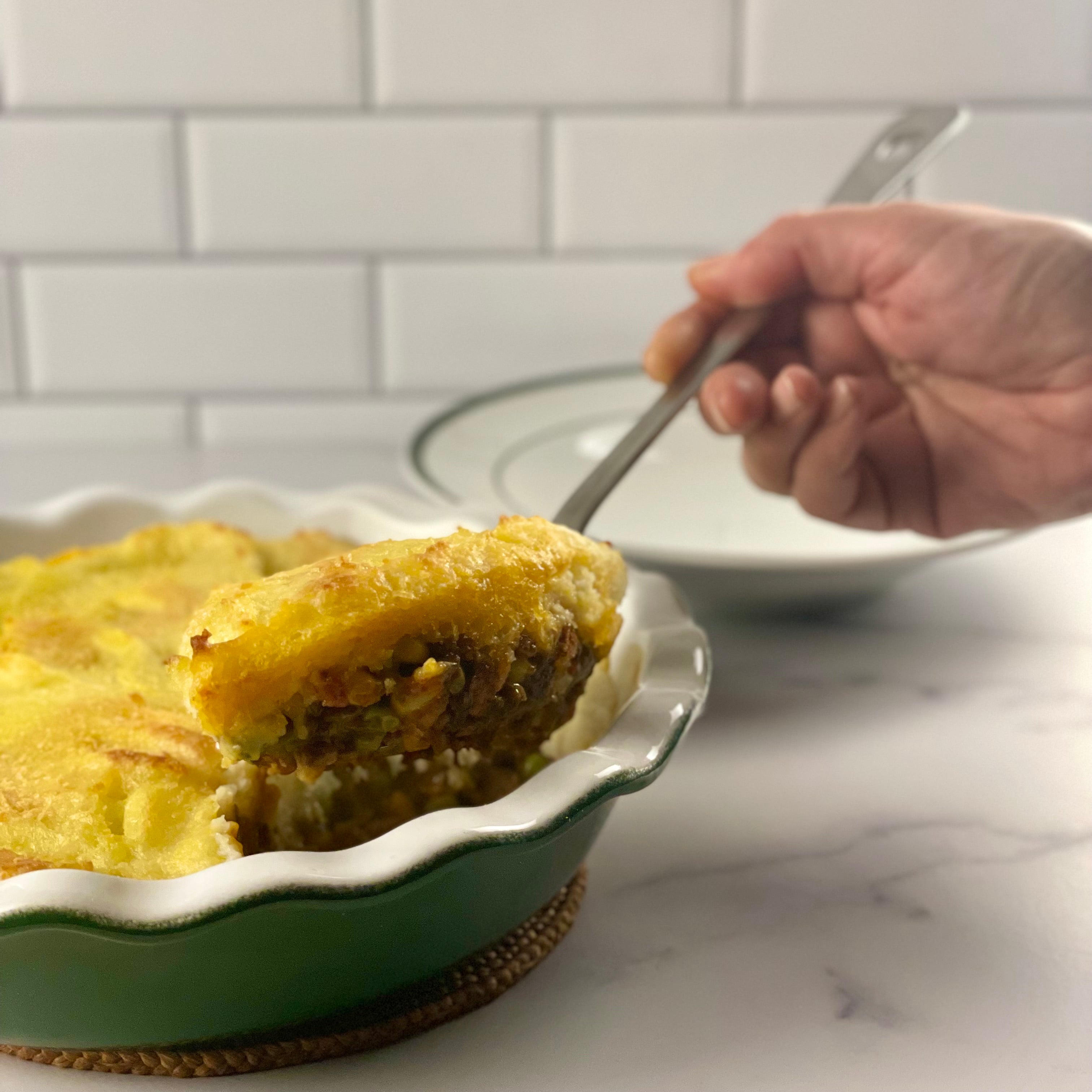 Close up image of Donald's hand as he scoops a generous serving of Irish cottage pie from an Emile Henry pie dish into a brasserie plate with a large stainless steel spoon.