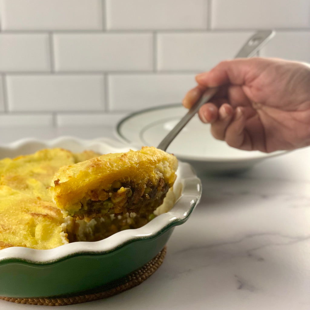Close up image of Donald's hand as he  scoops a generous serving of Irish cottage pie from an Emile Henry pie dish into a brasserie plate with a large stainless steel spoon.