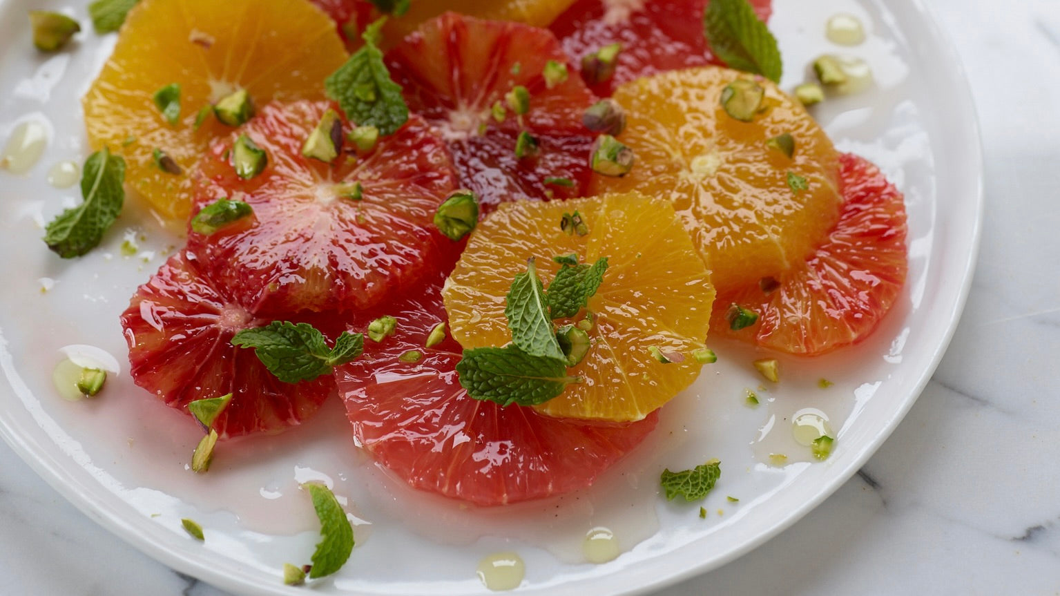 Luscious rounds of blood orange and mandarin orange slices on a flat round plate are festooned with chopped mint, chopped pistachios and a generous pour of Lucero Ascolano EVOO and Tagami Rosato Balsamic Vinegar