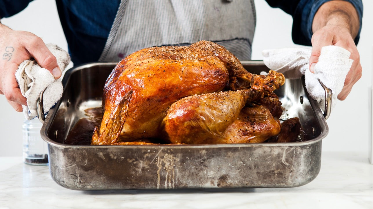 A man uses potholders to hold a roasting pan toward the view to show off a perfectly roasted whole turkey