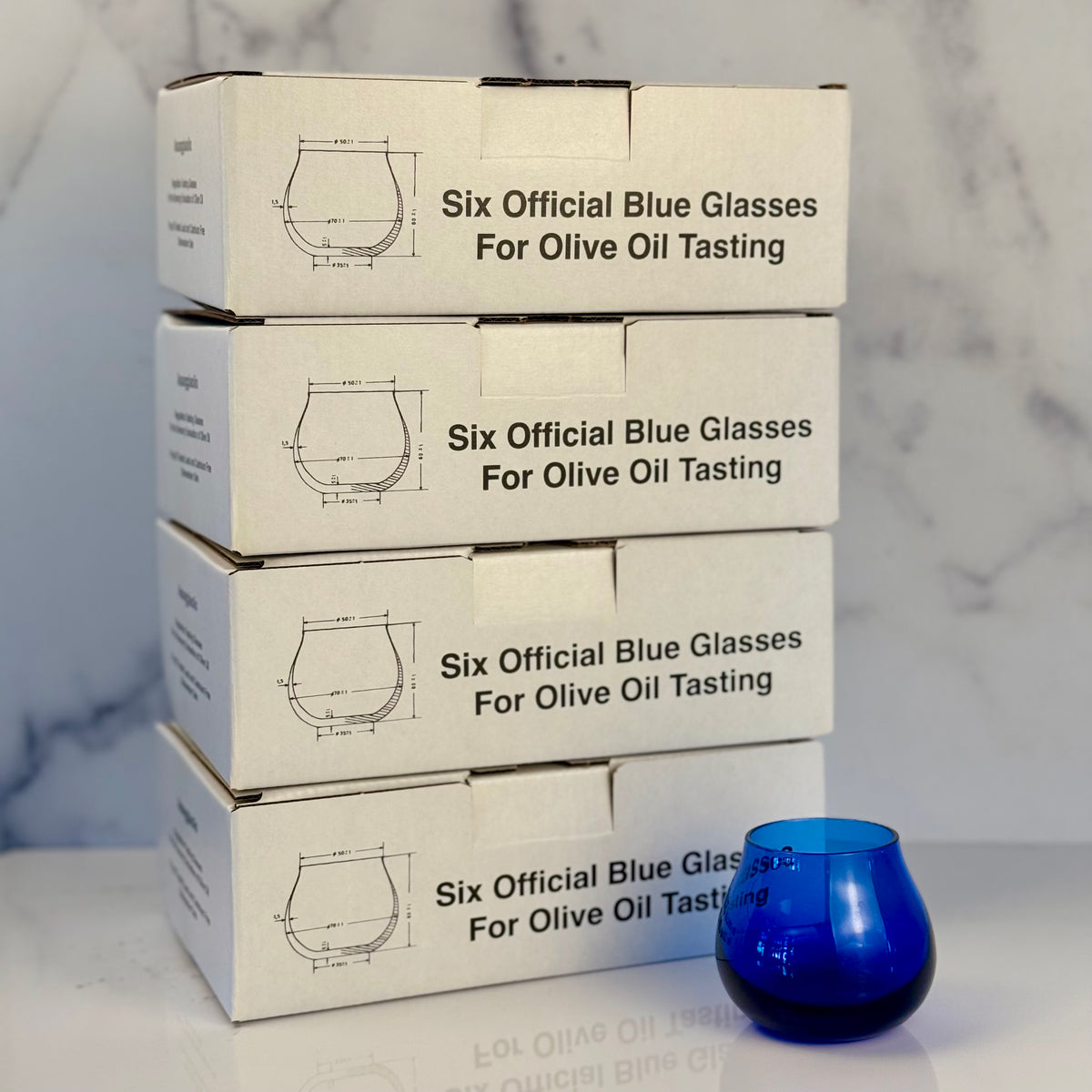 Four stacked white cardboard boxes labeled 'Six Official Blue Glasses For Olive Oil Tasting' on a white marble surface, with a single cobalt blue glass placed in front.
