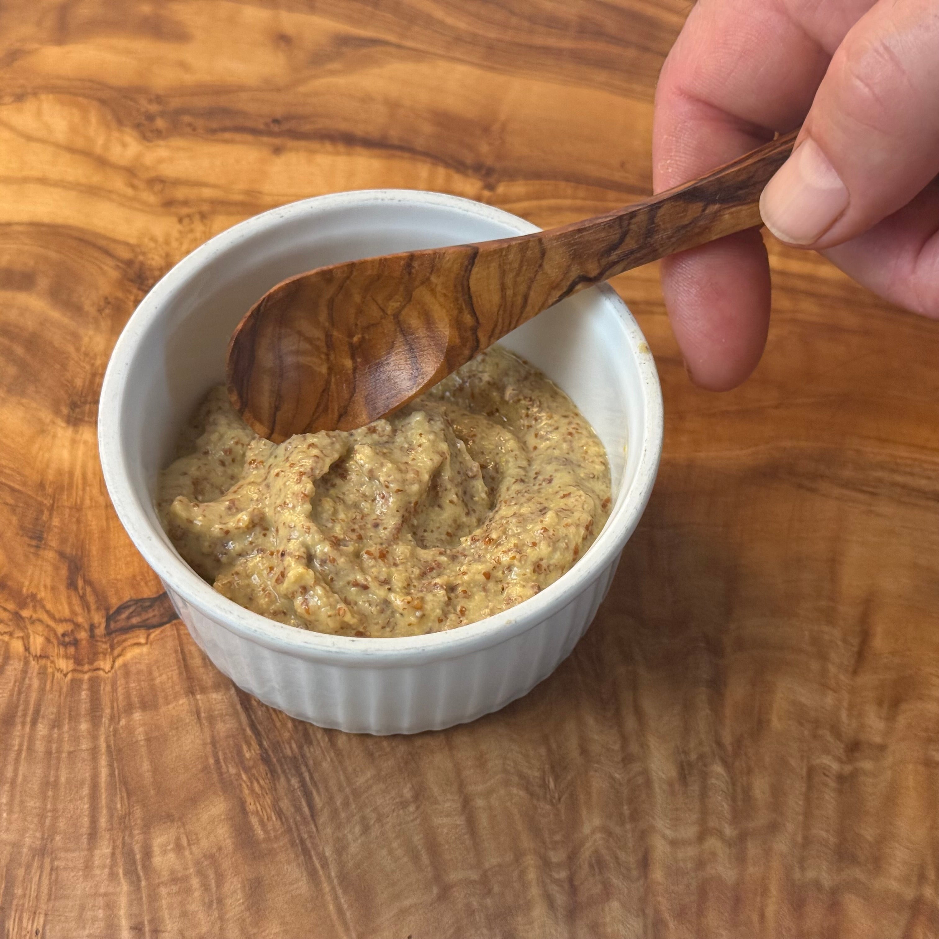 White bowl with mustard on a wooden surface, being stirred with a wooden spoon.