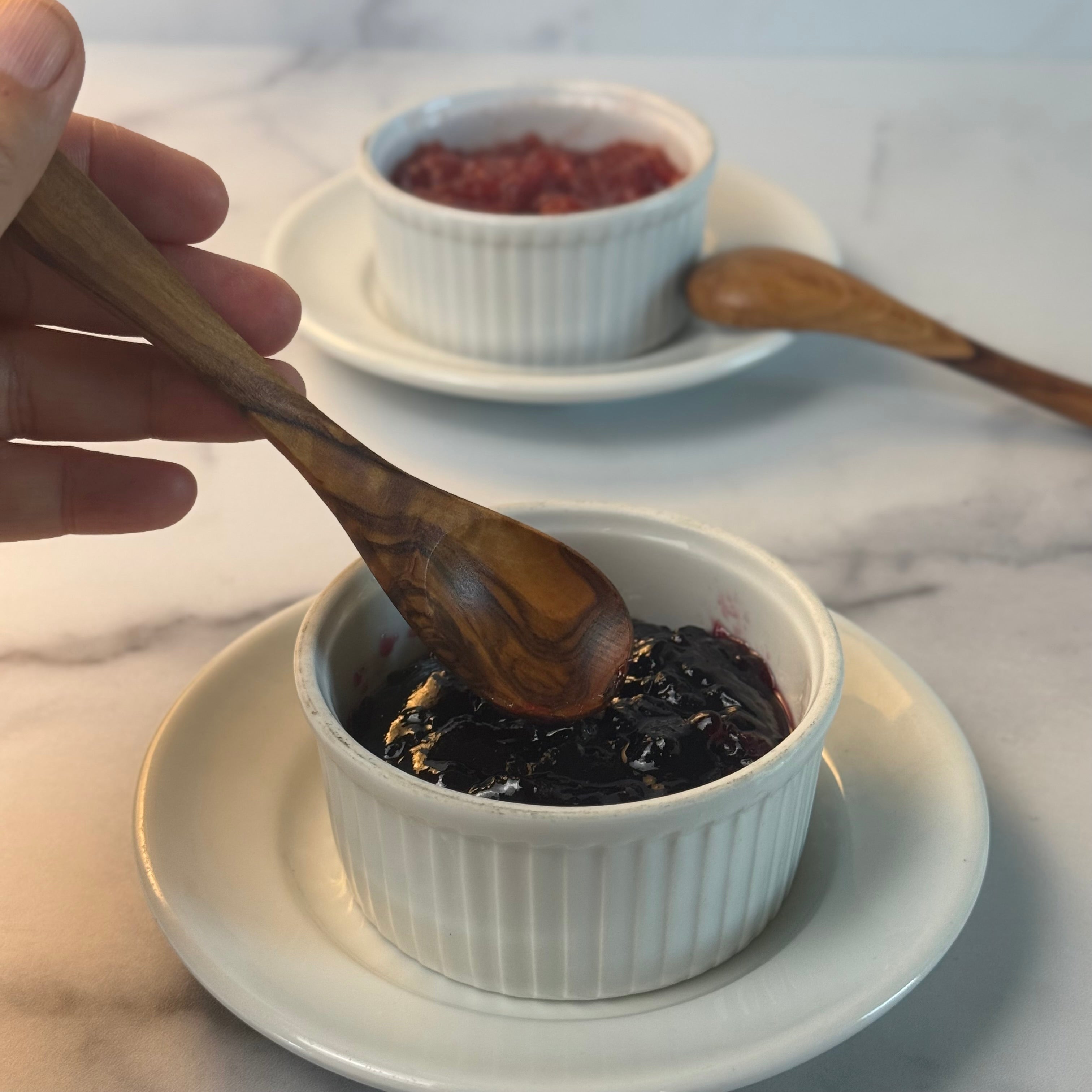 Hand using a wooden spoon to stir dark purple jam in a white ramekin on a marble surface.