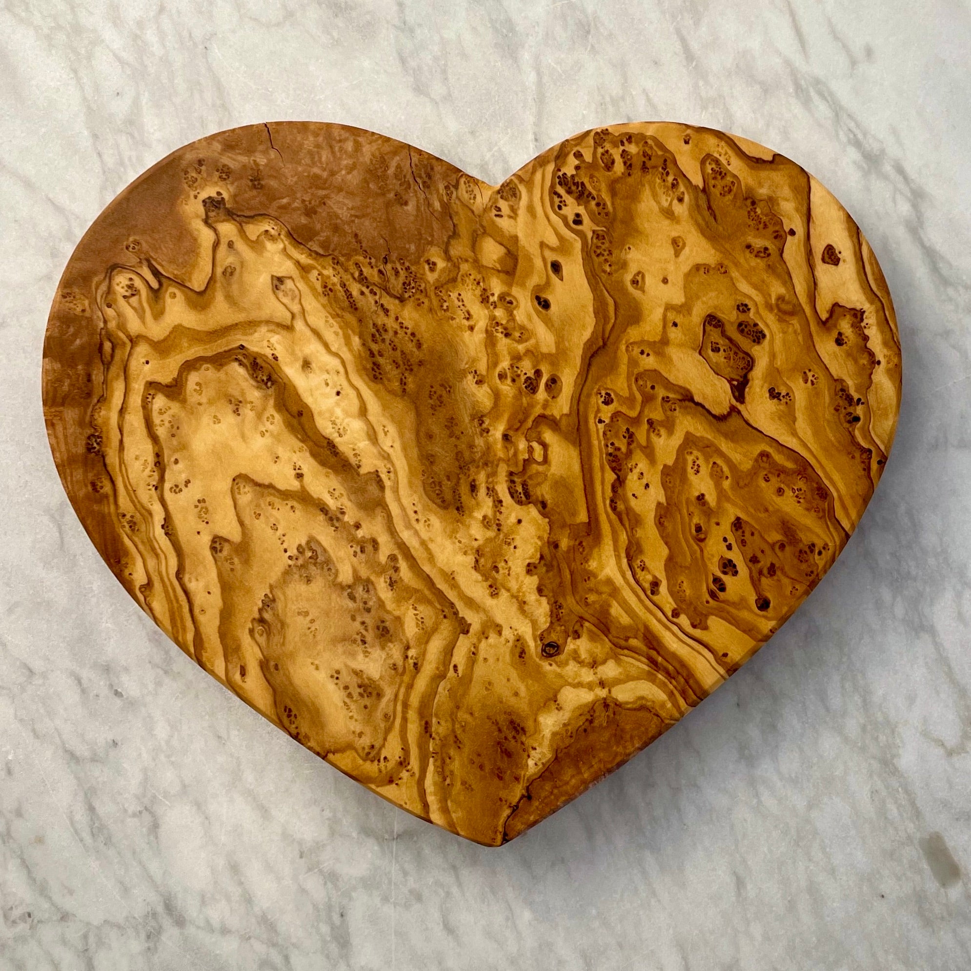 A highly figured olive wood board in a simple heart shape is shown on a marble counter