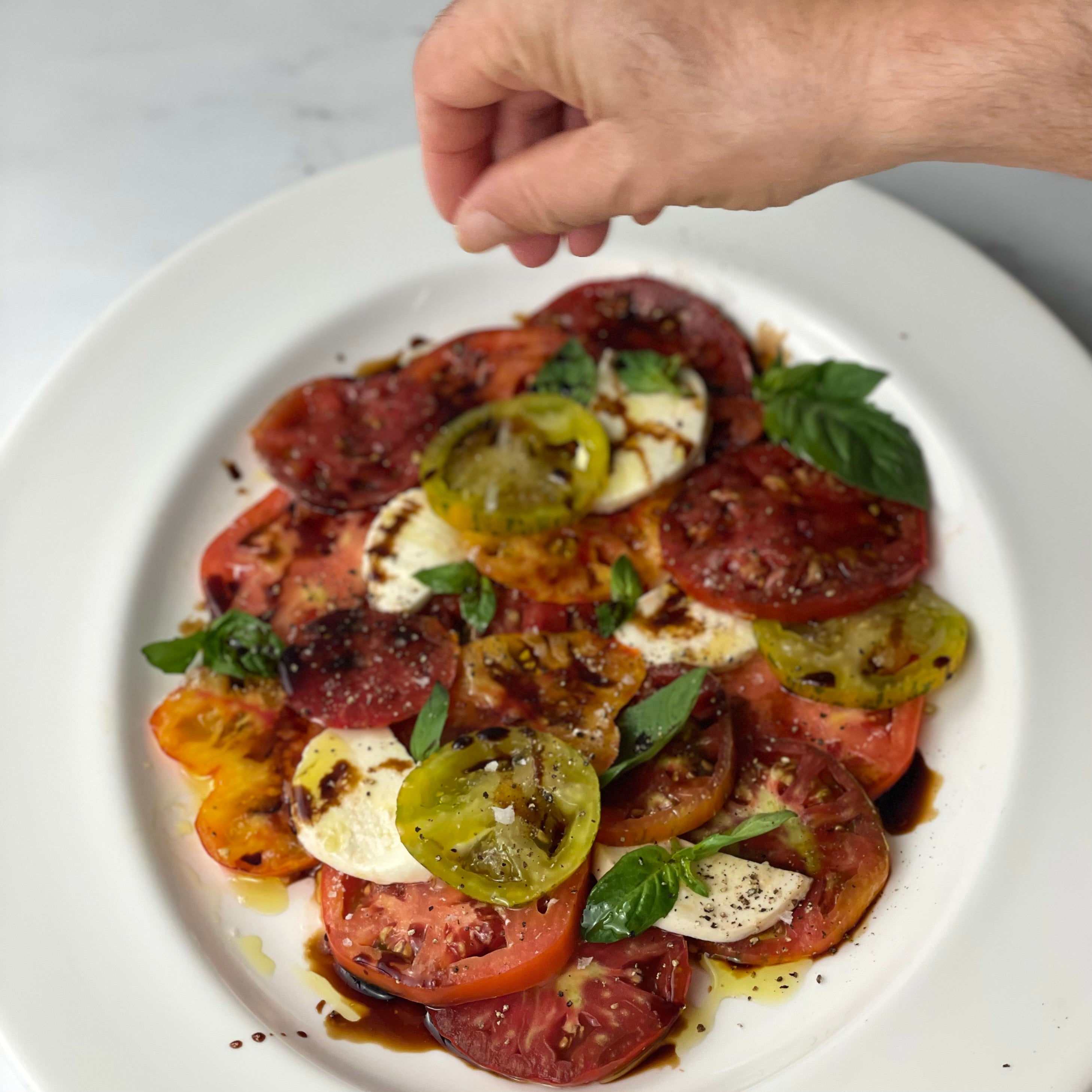 A close-up of a hand sprinkling seasoning over a white plate of caprese salad, featuring sliced red and green tomatoes, fresh basil leaves, and mozzarella cheese.