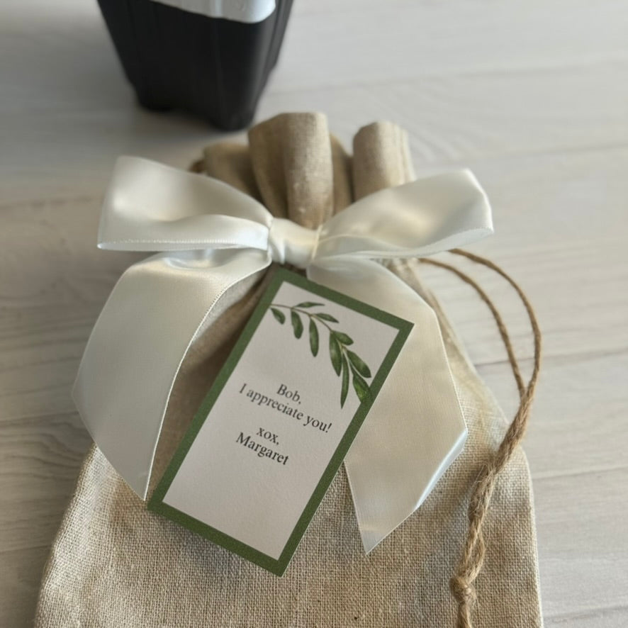 This is a close up image of the top of the standard natural linen burlap gift bags that are used at American Olive Farmer for our olive trees. Only the top half of the bag is shown so greater detail is seen with the soft texture of the fabric, the top is gathered, a white satin bow is placed on it, and  personalized gift card is shown on top. The base of a nursery pot is in the background nearly out of frame.
