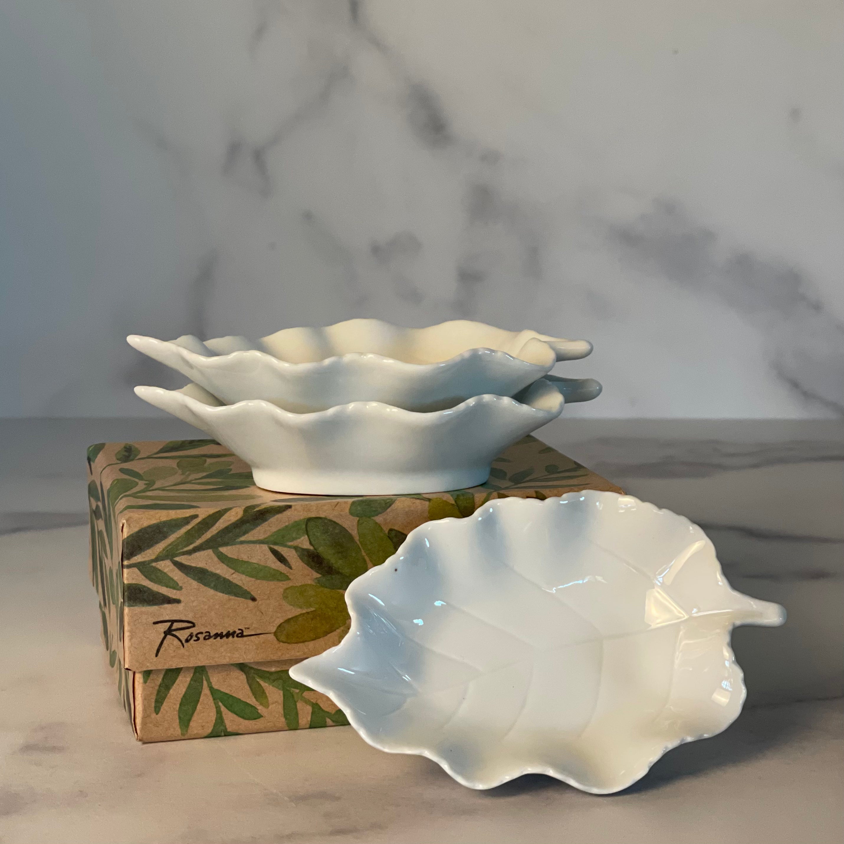 Three delicate white dipping bowls shaped like North American birch leaves are shown stacked, one facing out, on a decorative gift box. The dishes and box are on a marble counter.