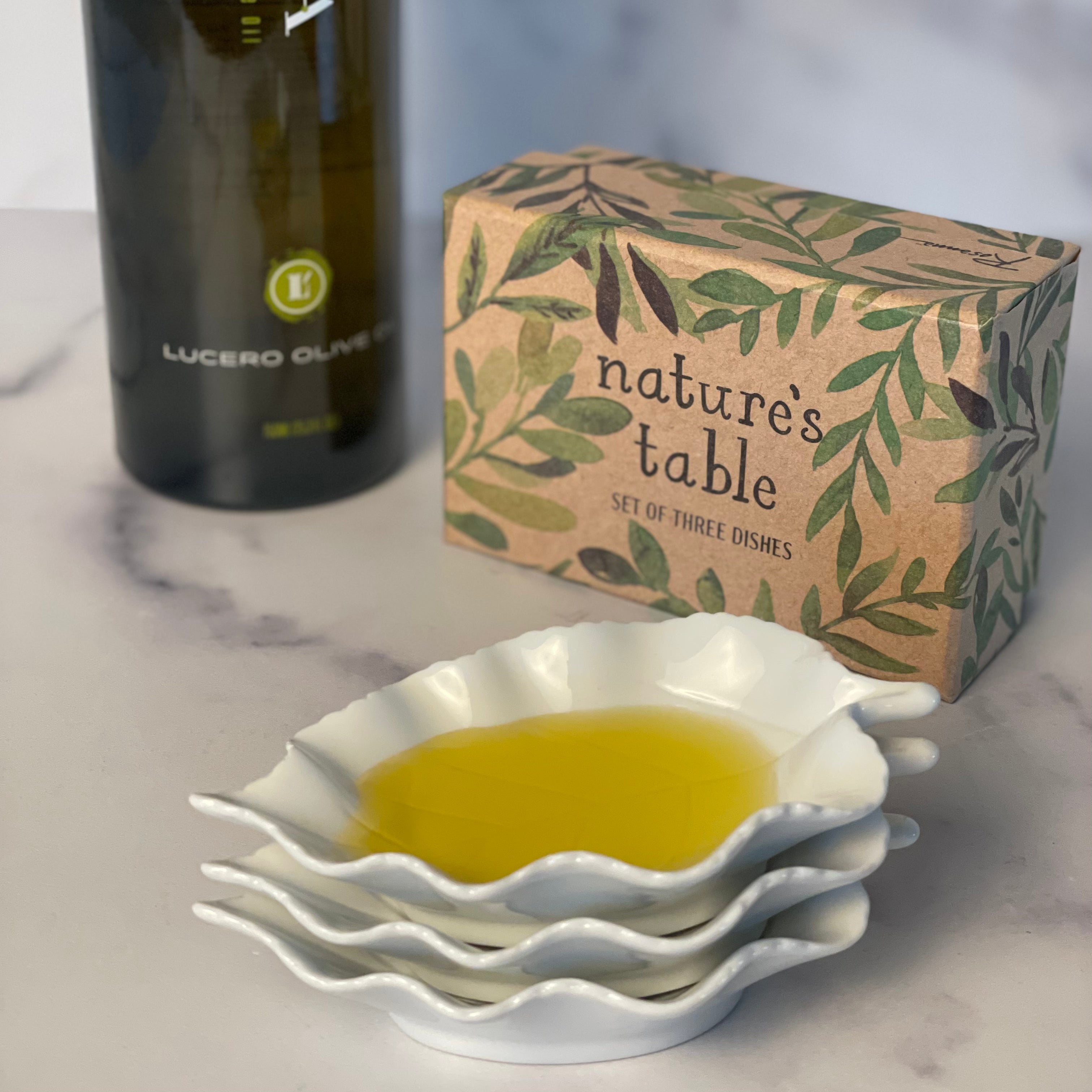 Three white porcelain dipping dishes shaped like birch leaves are shown stacked. The top one is filled with olive oil. There is a bottle of Lucero Olive Oil in the background next to a decorative gift box.