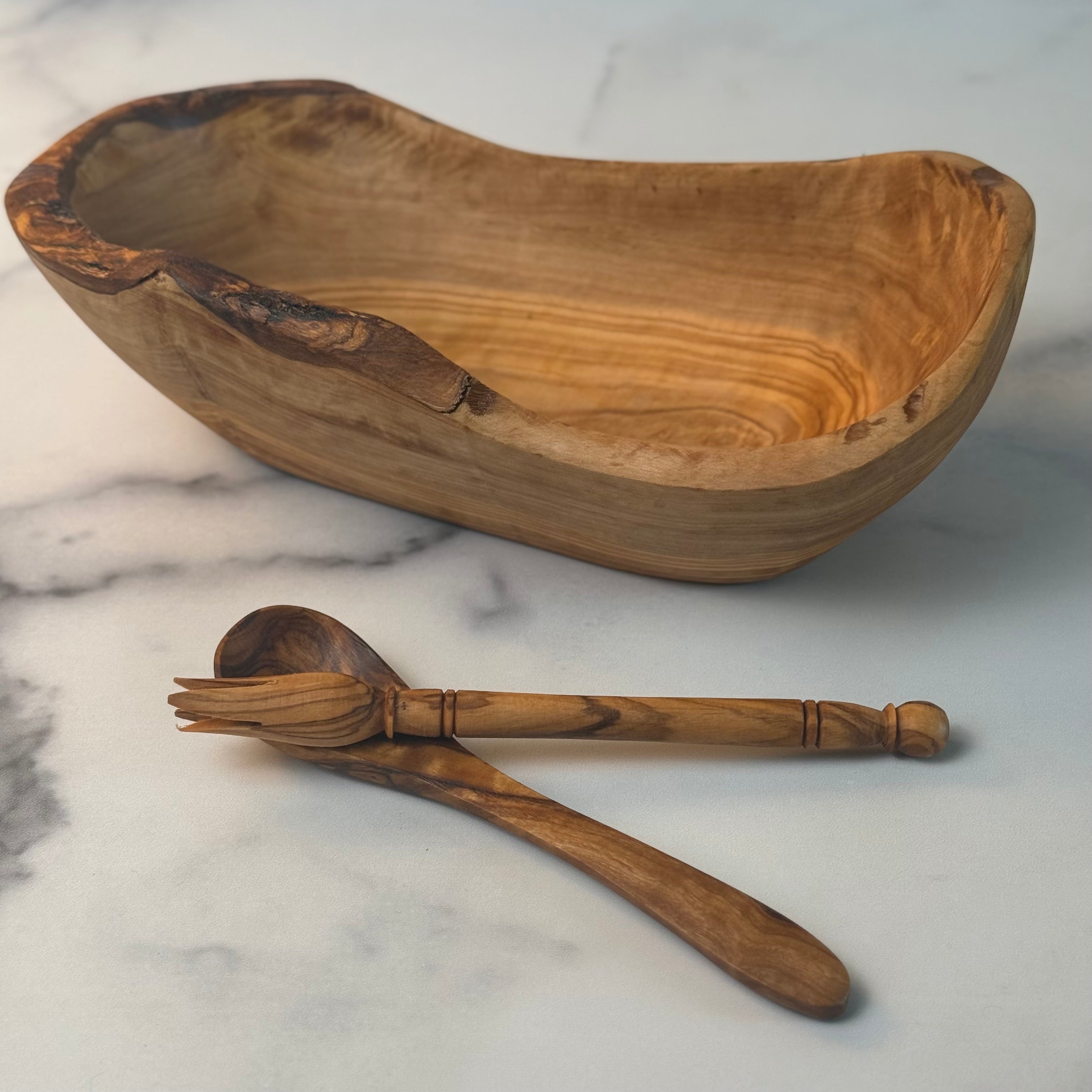 A rustic wooden bowl and set of matching olivewood utensils for serving olives on a marble surface