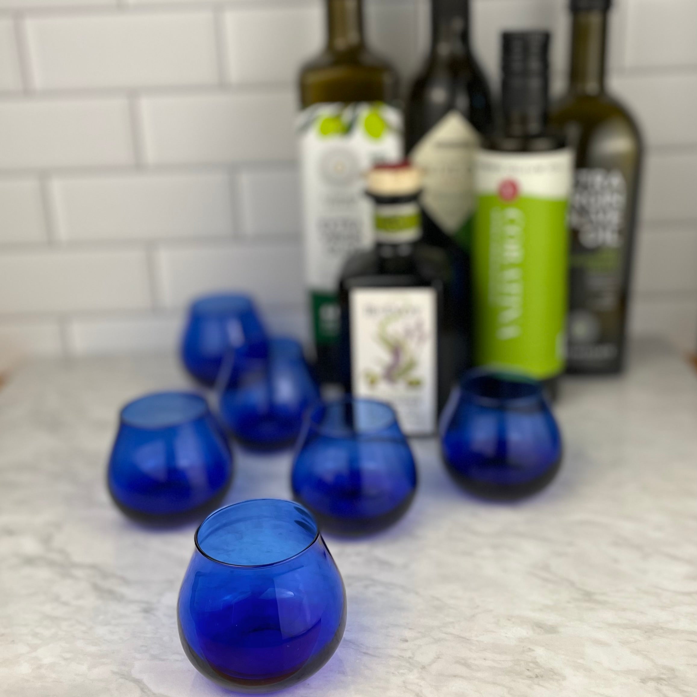 five olive oil bottles are shown out of focus in the background with five olive oil tasting glasses arranged nearby, also in the background. A single, in focus olive oil glass is highlighted in the front of the image, almost like an invitation to pick it up!