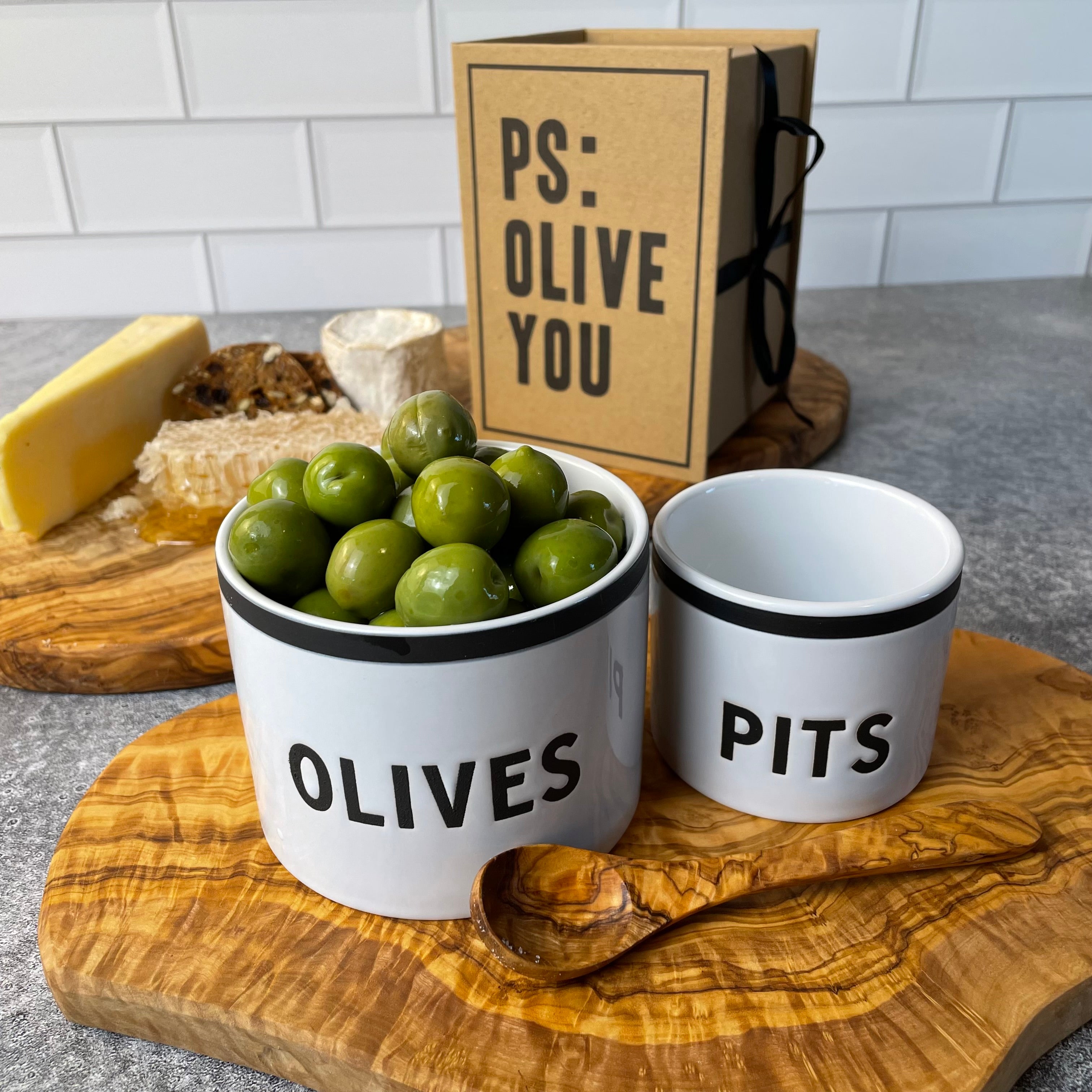 A set of two modern ceramic bowls marked OLIVES and PITS are shown on an olive wood board with an olive wood spoon. A charming cheese board with two types of cheese, honey comb, and artisan crackers are on another olive wood board, and the gift packaging for the OLIVE and PITS bowl is shown in the background.