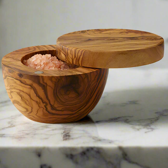 A round olive wood salt box is shown with the hinged lid swung open to reveal coarse grained pink Himalayan salt inside. The salt box sits on a dramatic white marble counter.