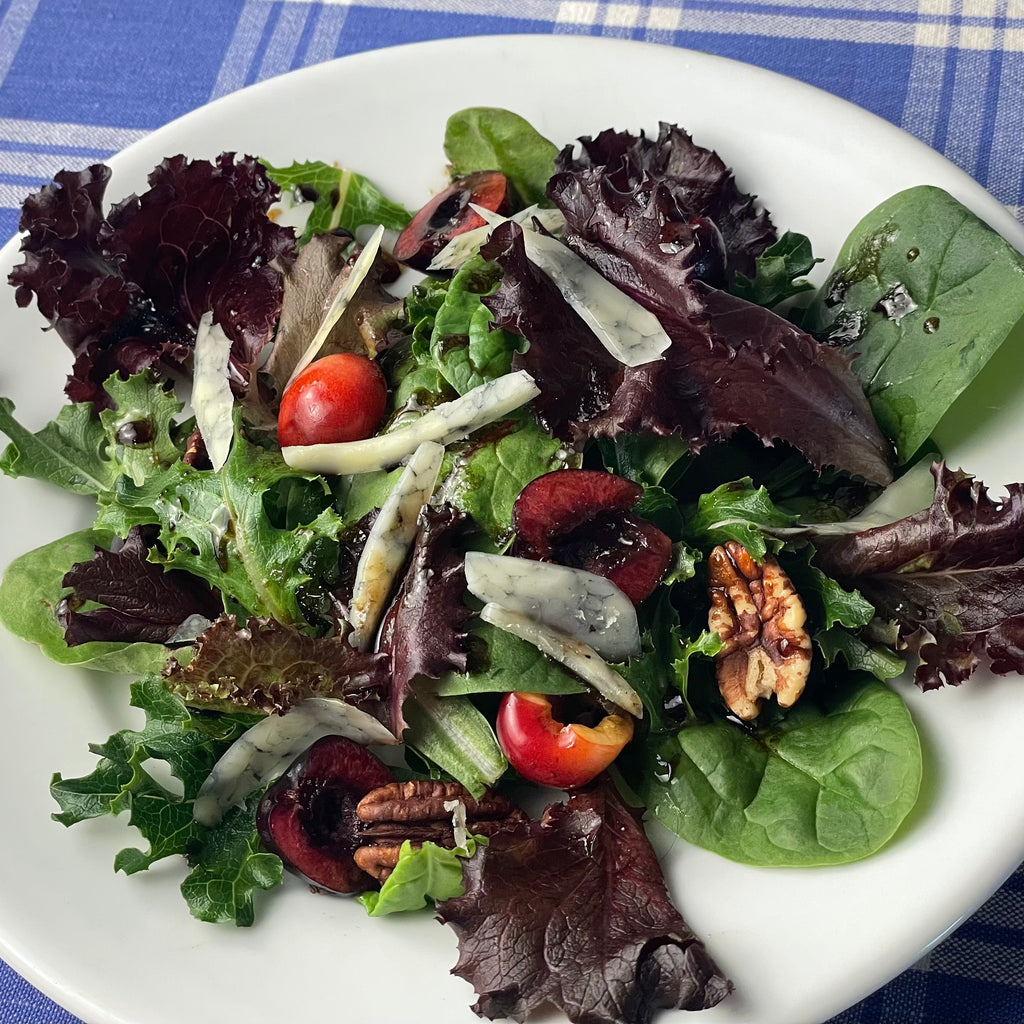 Mixed baby greens are adorned with fresh cherry halves, toasted pecans, and thin slices of blue cheese on a small French porcelain plate.