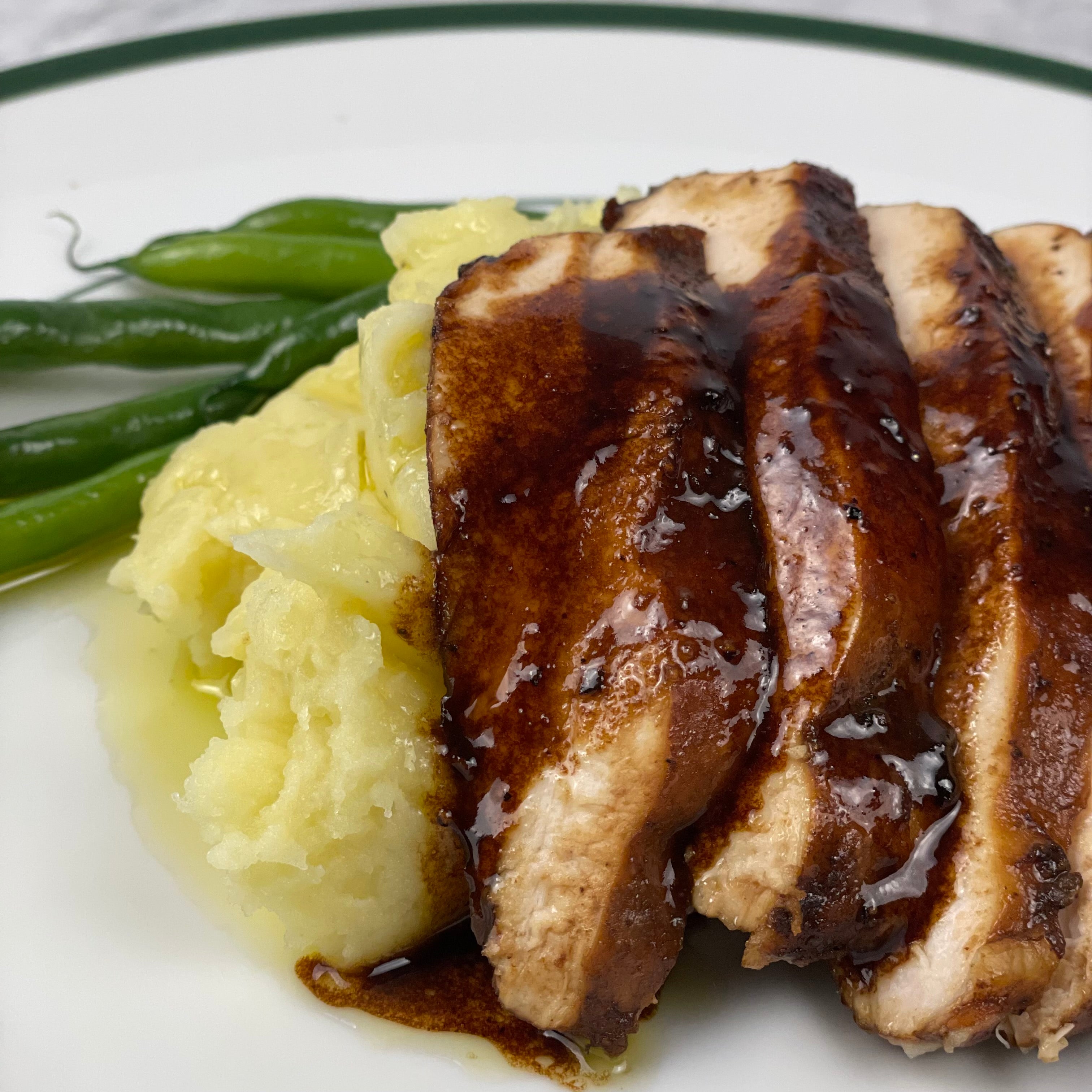 Close up image of sliced chicken with balsamic butter sauce on top of mashed potatoes with green beans in background.