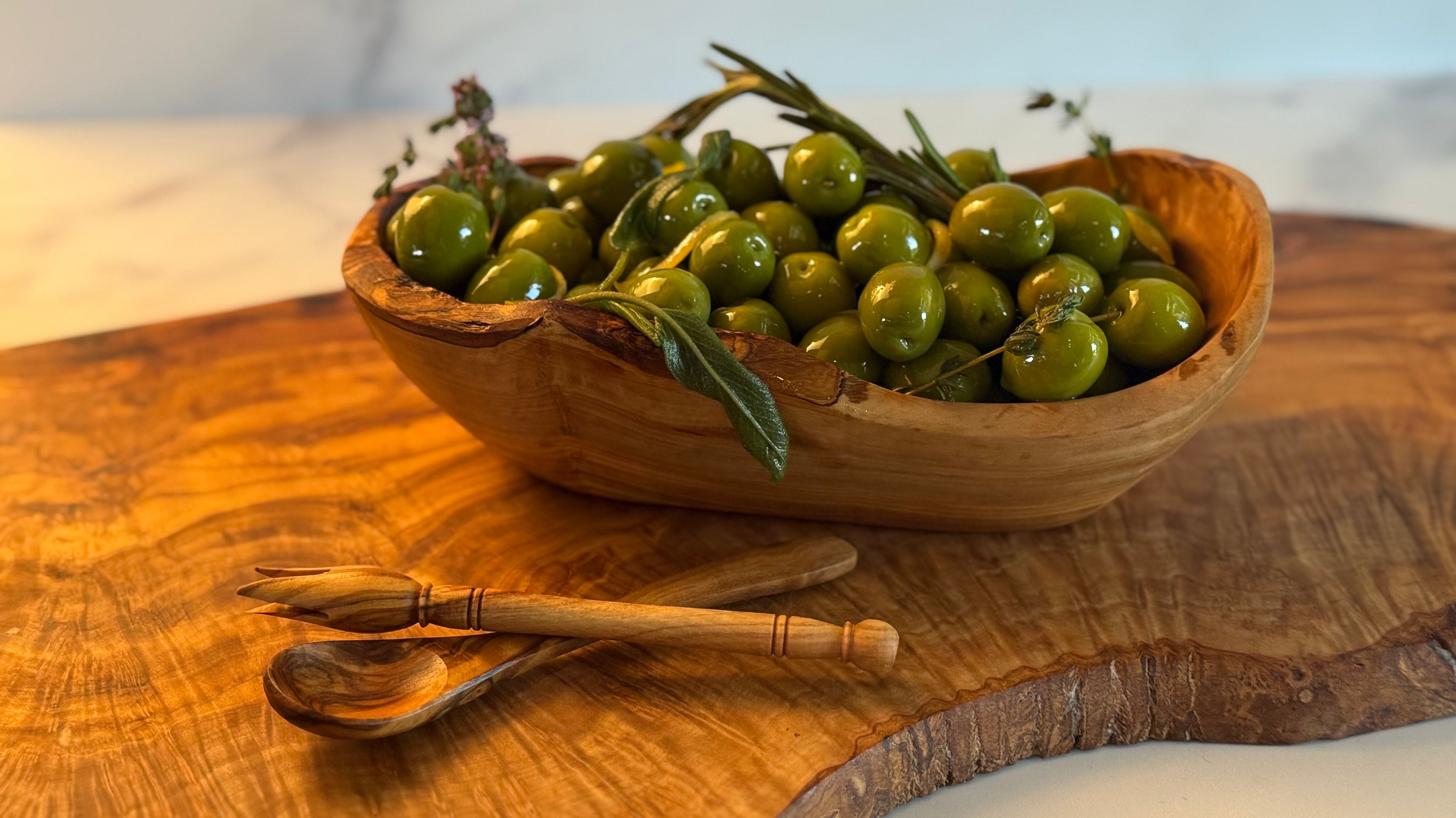 Olivewood serving boat is shown full of olives. There are two serving utensils nearby, a spoon and olive fork, and all three pieces are shown on an olivewood board.
