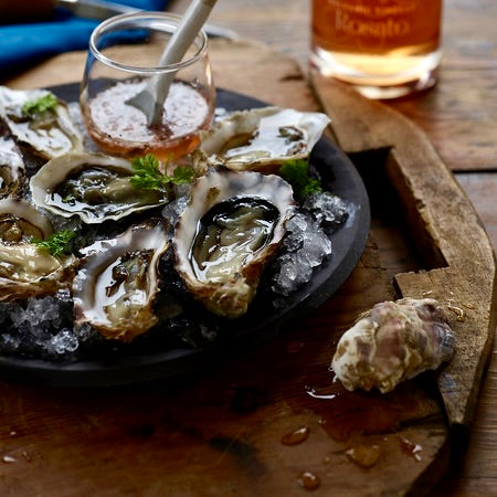 Oysters on the half shell are arranged around a transparent vessel showing a mignonette sauce inside. The sauce appears to already be sprinkled on the oysters.