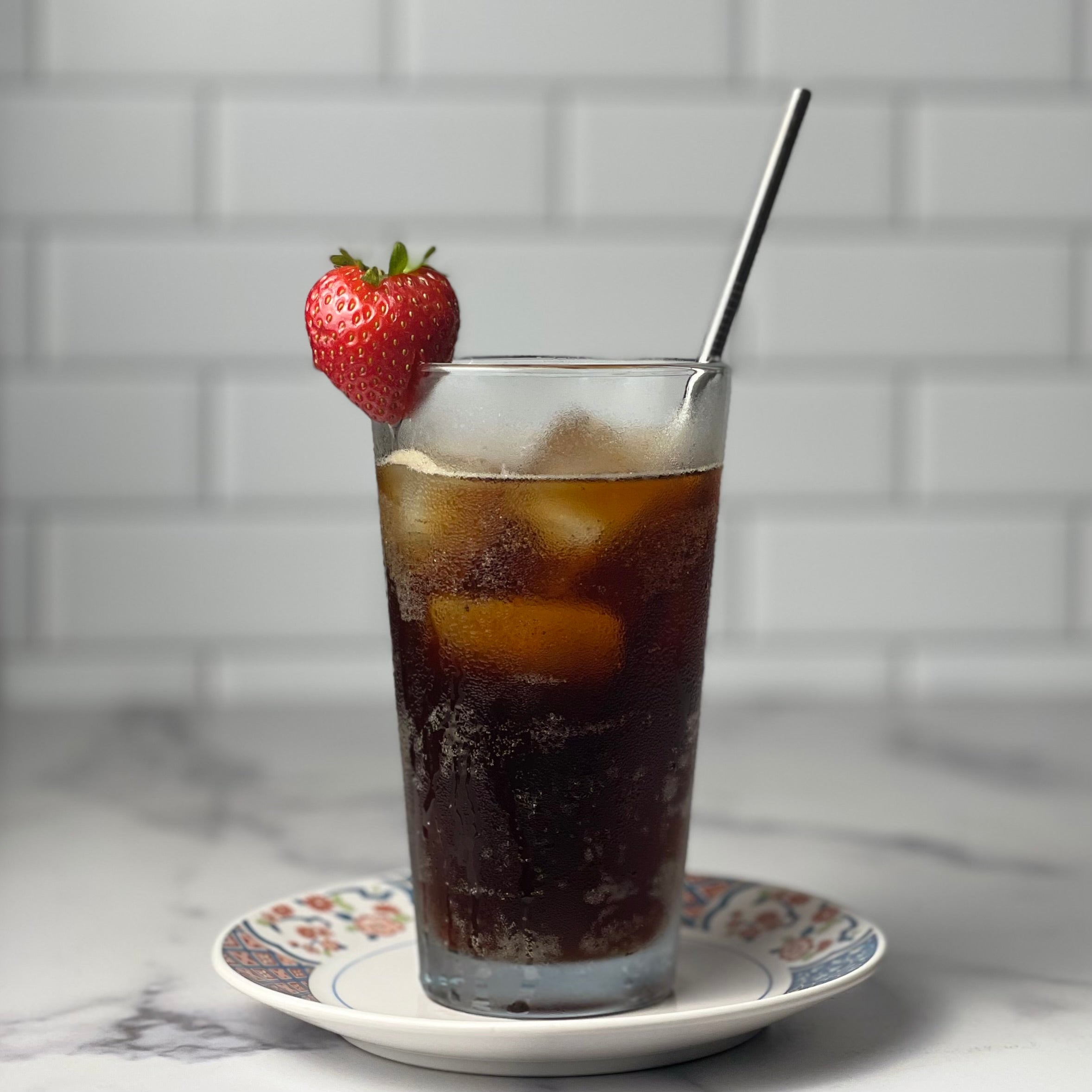 A pint glass is filled with ice and a refreshing beverage with bubbles. A strawberry is stuck to the rim of the glass, and a stainless steel straw sticks out at a jaunty angle. Scene is in a marble and tile kitchen.