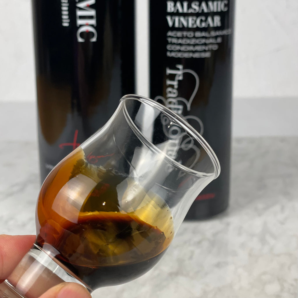 Close up image of viscous balsamic vinegar in an official Italian tasting glass with both small and large bottles of vinegar in the background