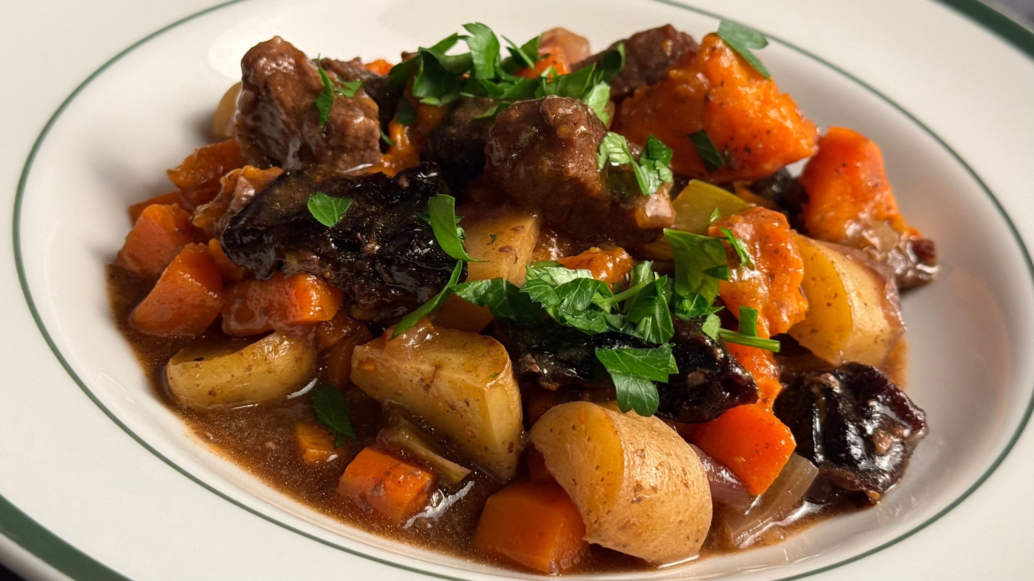 A French porcelain soup plate is filled with luscious roasted beef and stewed prunes, sweet potato, new potatoes, and carrots in a savory sauce.