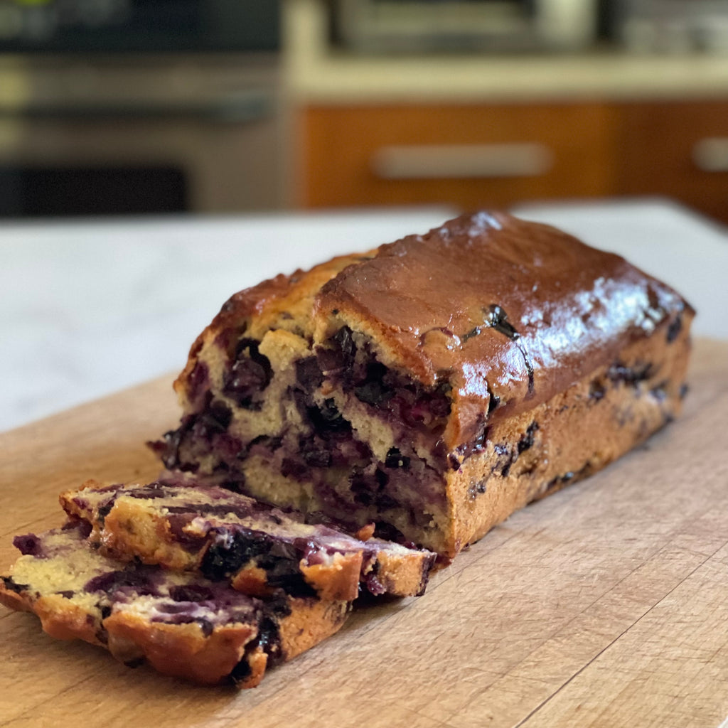 A freshly sliced loaf of blueberry olive oil cake sits on a  wooden cutting board. Two slices reveal a high percentage blueberries inside.