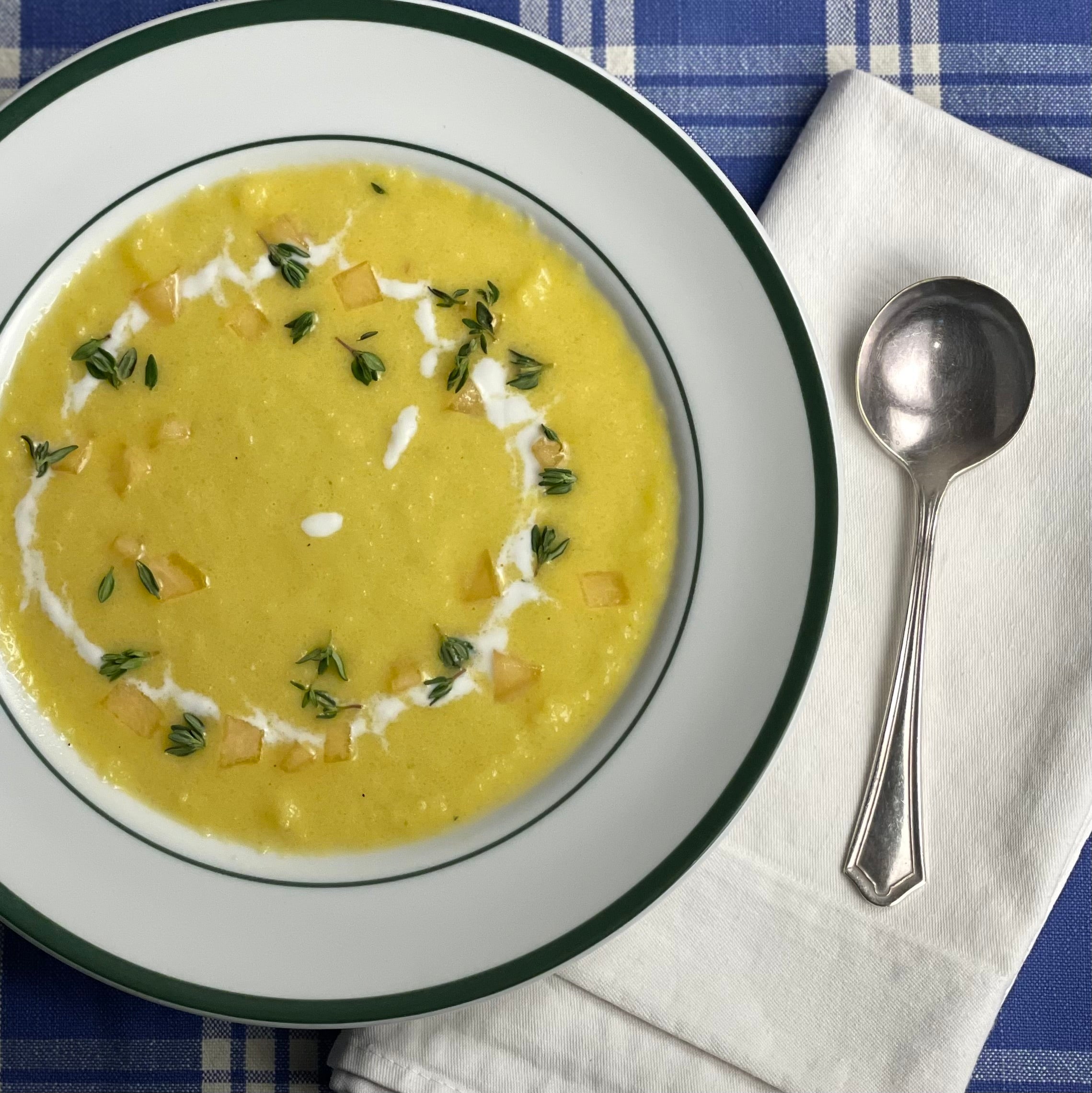 An 8" European soup plate holds a smooth soup with small bits of melon, a swirl of yogurt, and sprinkle of fresh thyme.  A freshly pressed cotton napkin and vintage silver soup spoon are shown just to the left