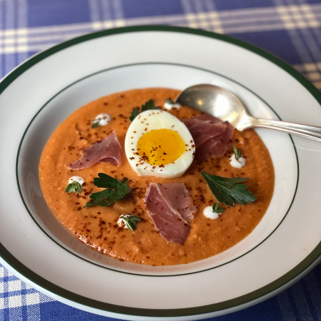 A classic soup plate is filled with creamy tomato soup embellished with half an egg, slices of ham, sour cream, and herbs artfully arranged and sprinkled with chili flakes