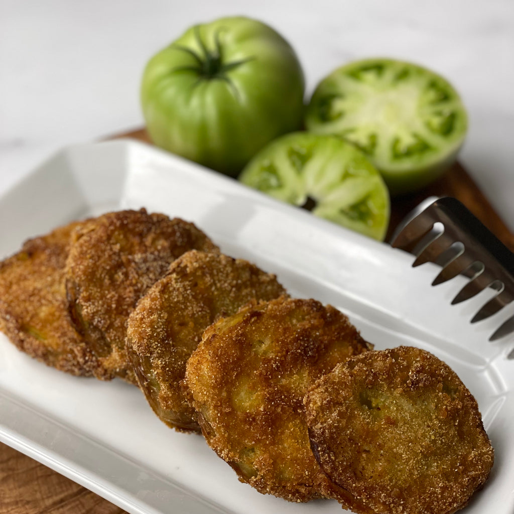 Five crispy rounds of fried green tomatoes are arrayed on a chunky French porcelain plate. One whole and one sliced uncooked green tomato are shown in the background.