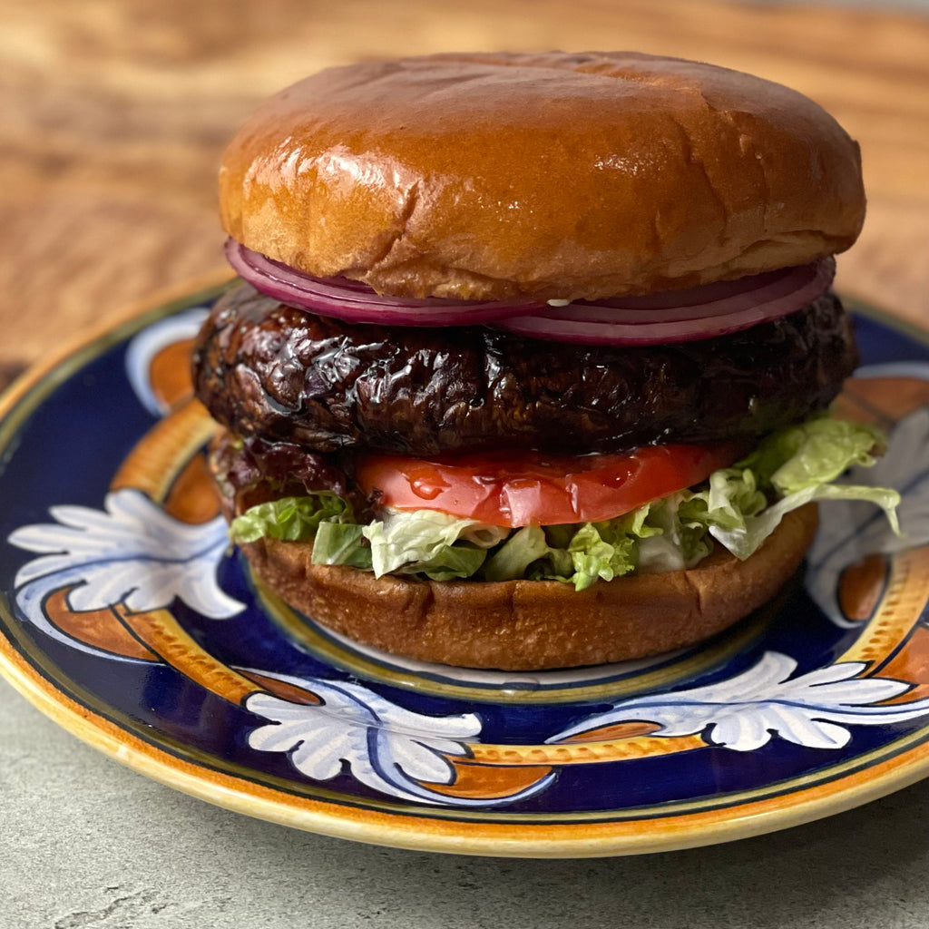 A juicy grilled portobello mushroom is shown enclosed in a hamburger bun with layers of romaine lettuce, heirloom tomato, and vinegar marinated onion on a 20 cm Deruta-style plate.