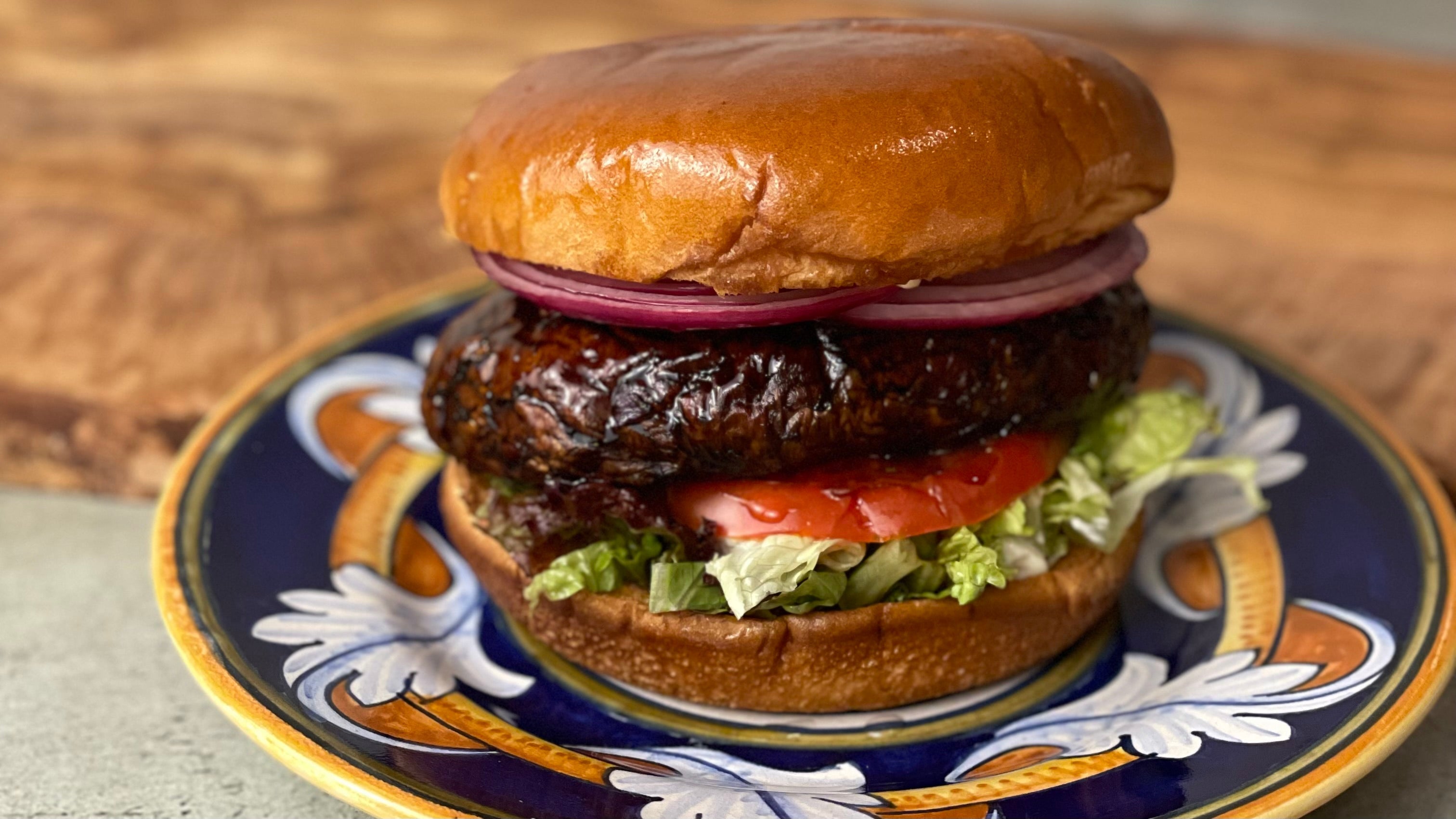 A succulent looking whole glazed portabello mushroom is the highlight in a burger shown with red onions, tomato, and lettuce on a brioche style bun. The burger is on a colorful Italian ceramic plate that complements the colors of the burger.