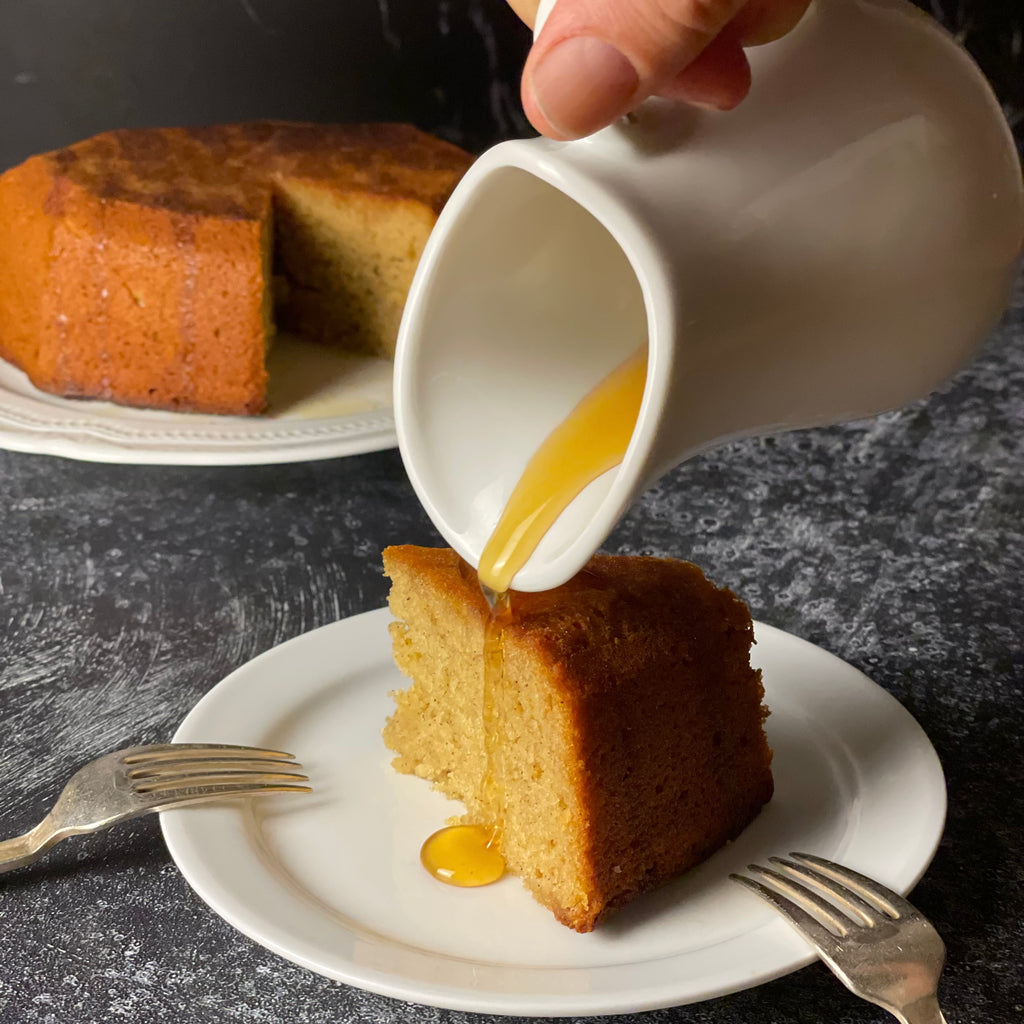 Warm honey and rosemary syrup is poured over a moist slice of apple honey cake with two forks. The rest of the cake is in the background