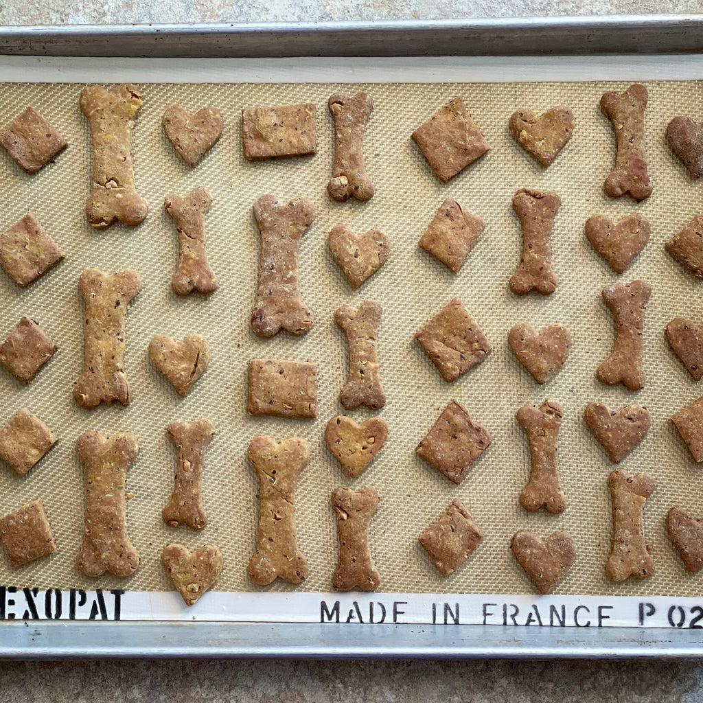 A professional weight half baking sheet is filled with small dog biscuits shaped like hearts, diamonds, squares, and "dog bones" rest on a silicone "exopat".