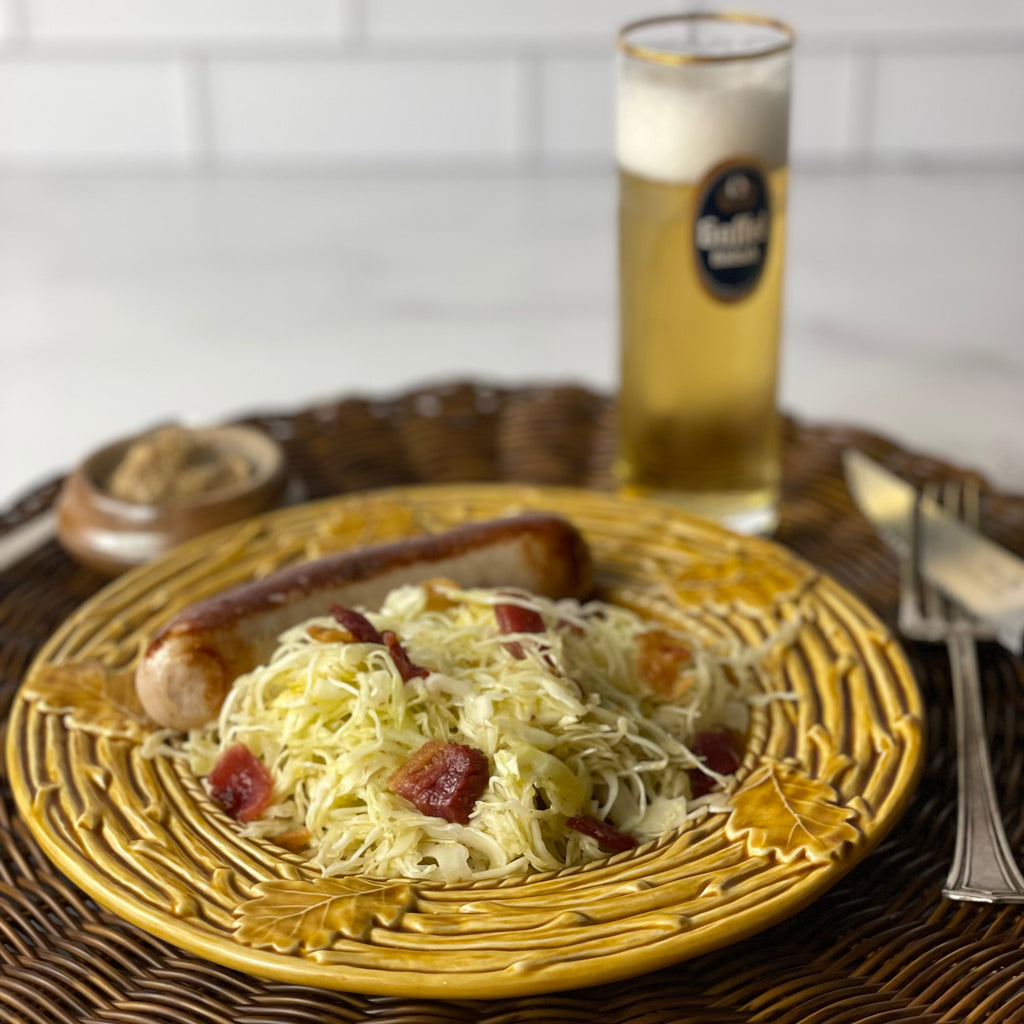 A small plate holds a generous portion of fresh cabbage and bacon salad with a white sausage in the back. A small pot of mustard and glass of beer with a good head in a frosty glass are ready to be enjoyed with the meal.