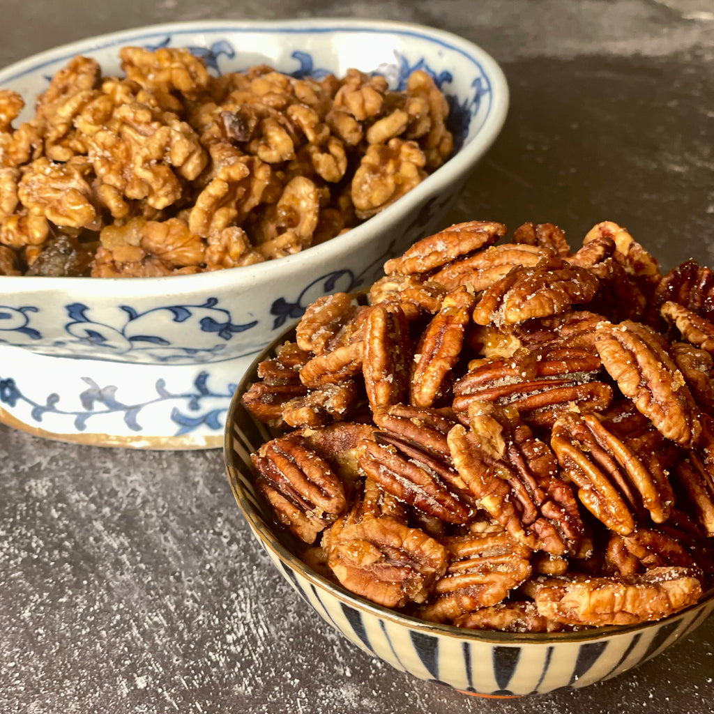 Lightly sugared fire dried walnuts in a blue and white Chinese porcelain pedestal bowl and lightly sugared fire sugared fire dried pecans in a blue and white Japanese porcelain rice bowl are shown close up on a plain background.