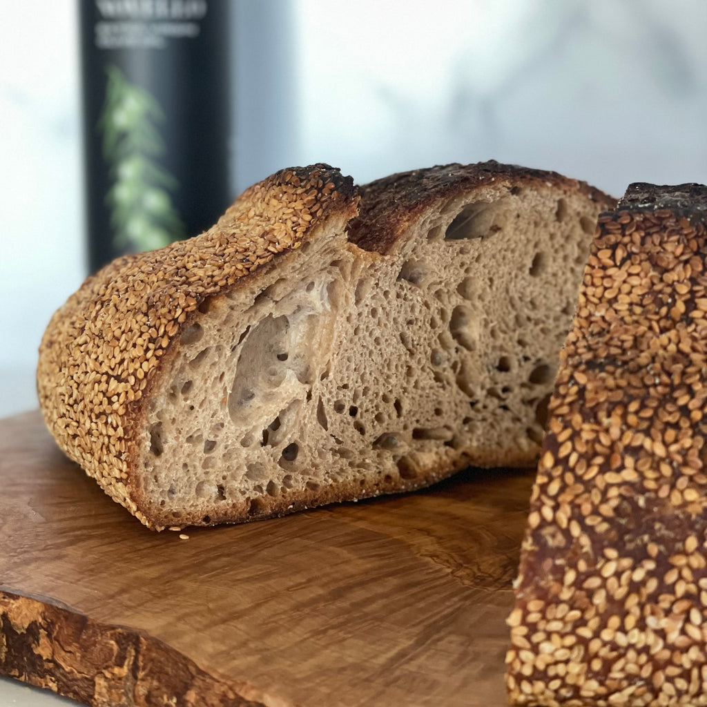 A chewy and dense hunk of Tartine style loaf made with 50/50 whole and white flour on an olivewood board. The loaf is opened and the outside is covered with sesame seeds. Olio Novello bottle is in the background.