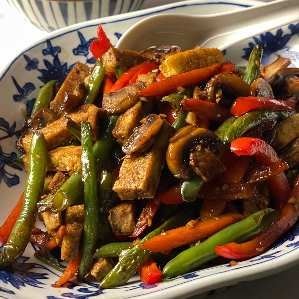 A vintage Chinese porcelain bowl, footed in a rounded diamond shape, holds a vegetarian stir-fry with green beans, red bell pepper, mushrooms, and crispy tofu batons in a glossy sauce.