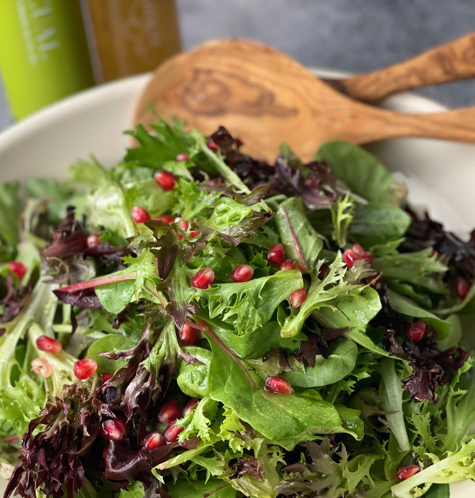 Mixed green salad with pomegranate seeds shown in a wide ceramic bowl with olivewood salad servers