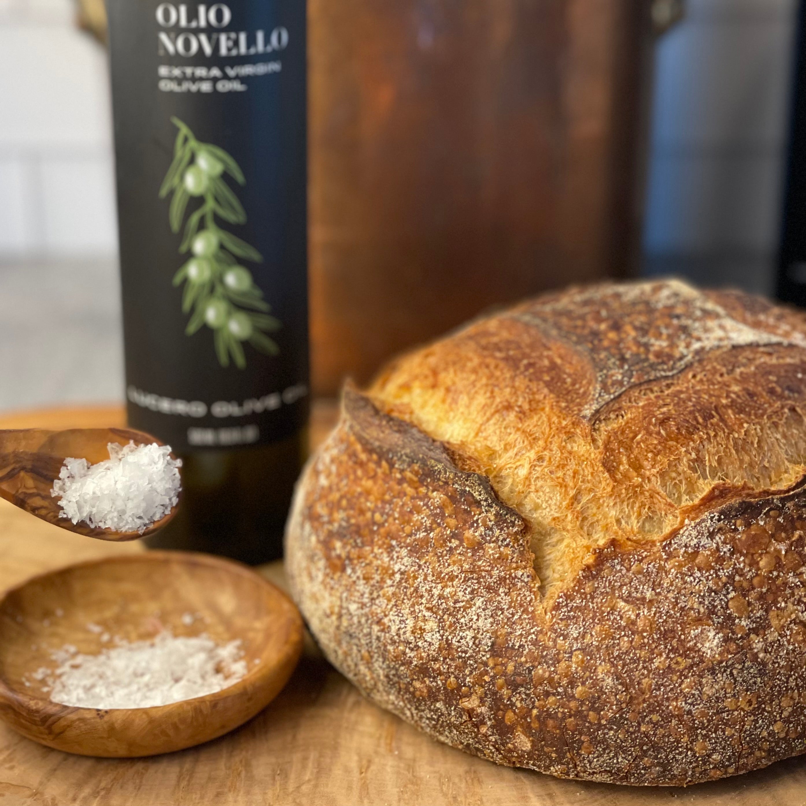 A crusty round country loaf sits on a board near Olio Novello and salt, waiting to be opened.