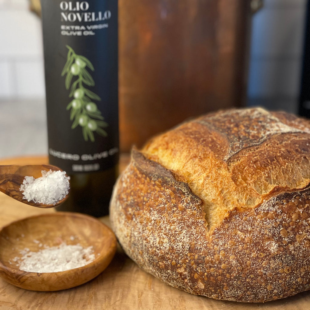 A crusty round country loaf sits on a board near Olio Novello and salt, waiting to be opened.