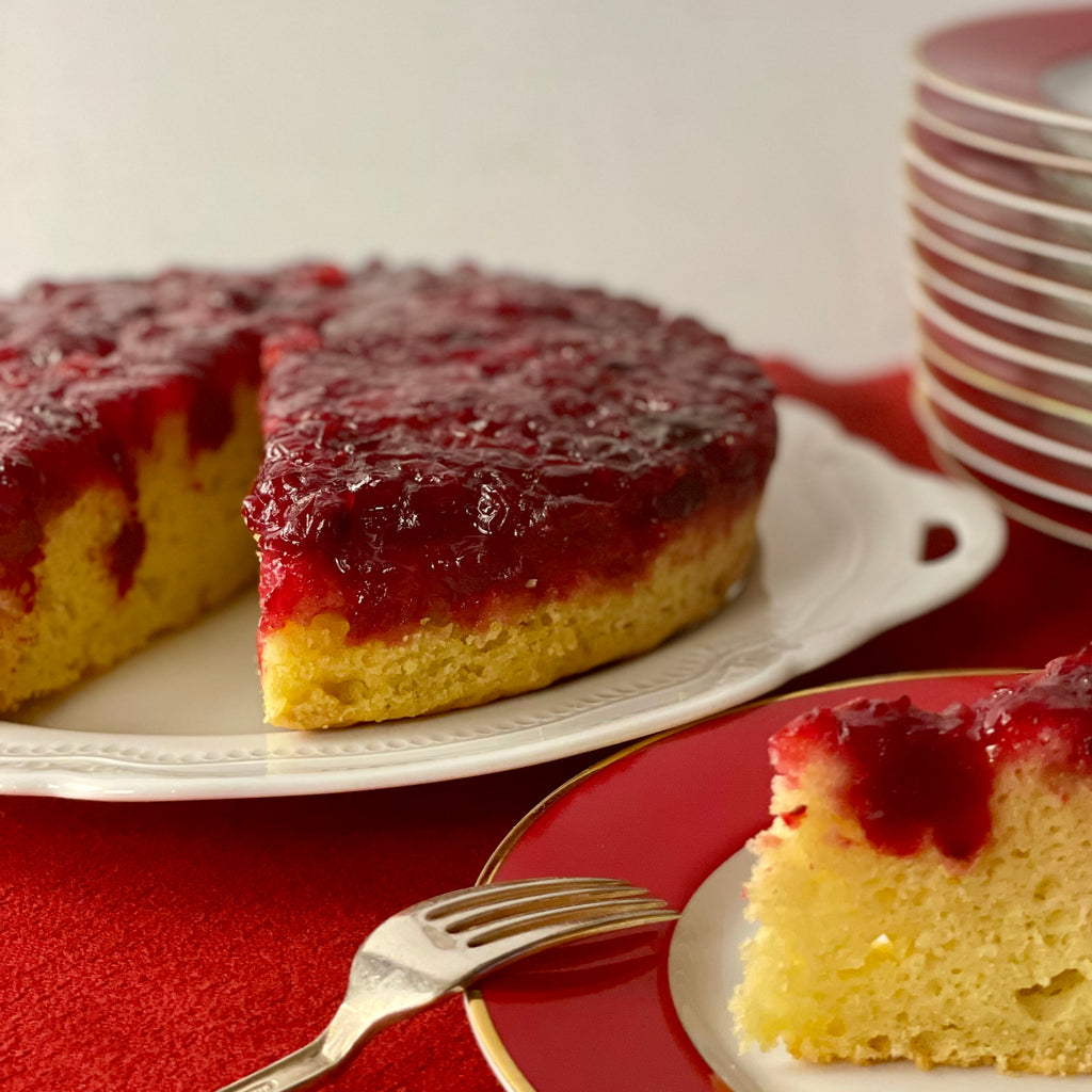 A 9" cake with cranberries has a single slice cut out and on a plate in the foreground.  A stack of dessert plates is stack in the background.