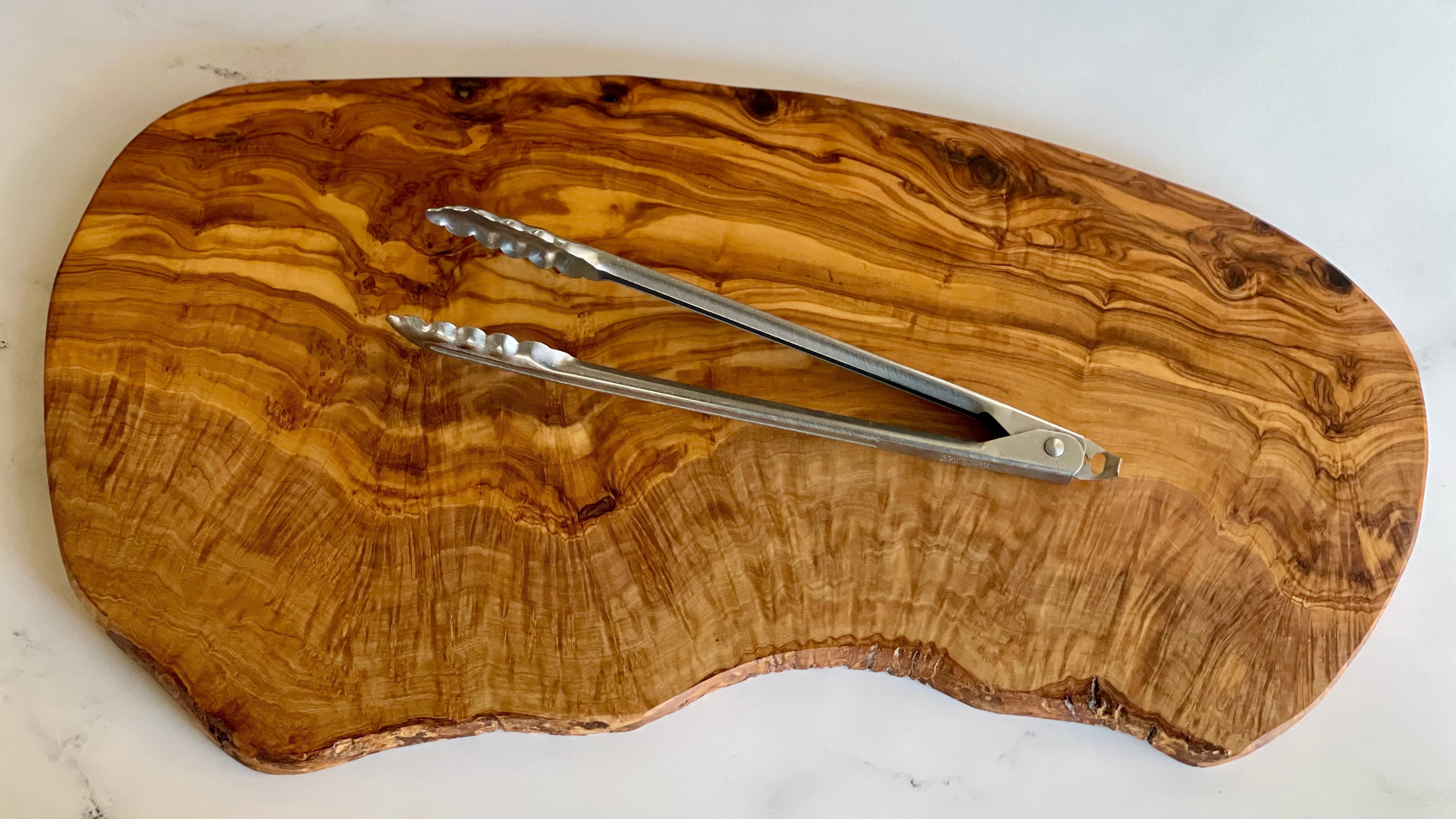 A large olive wood board shows striking markings and a undulating "live edge". A 12" pair of kitchen tongs are placed on top for scale.
