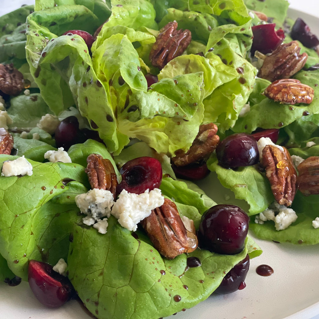 Mixed baby greens are adorned with fresh cherry halves, toasted pecans, and thin slices of blue cheese on a small French porcelain plate.