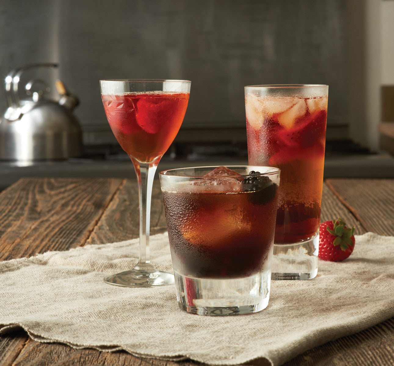 Three Fruity Balsamic Vinegar cocktails shown in a casual kitchen. One in a stemmed glass, one one a double old fashioned glass, and one in a highball glass.