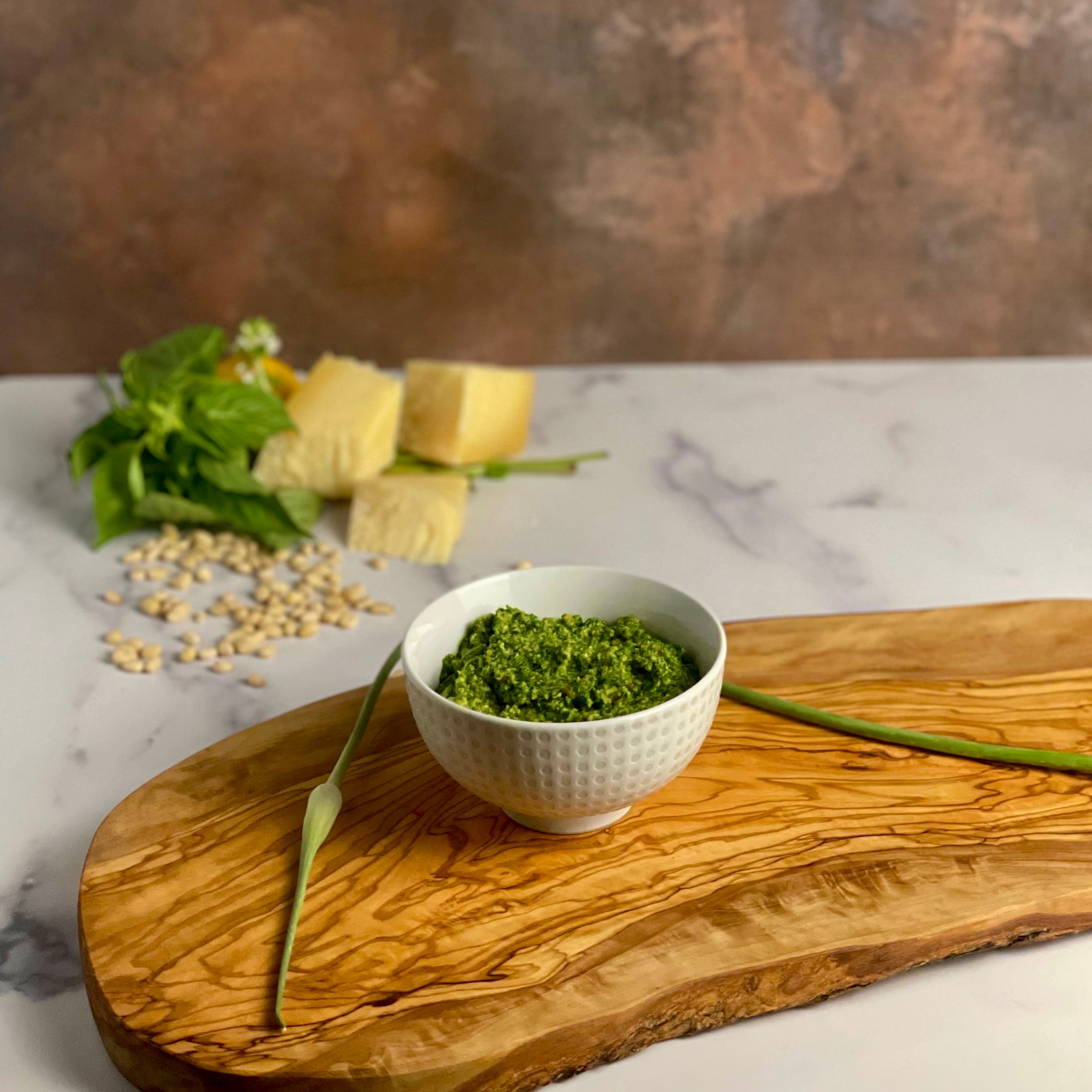 A small bowl of fresh pesto sits on an olivewood board in a marble and copper kitchen. A bunch of fresh basil, large chunks of parmigiano reggiano, and a scattering of pine nuts are shown out of focus in the background