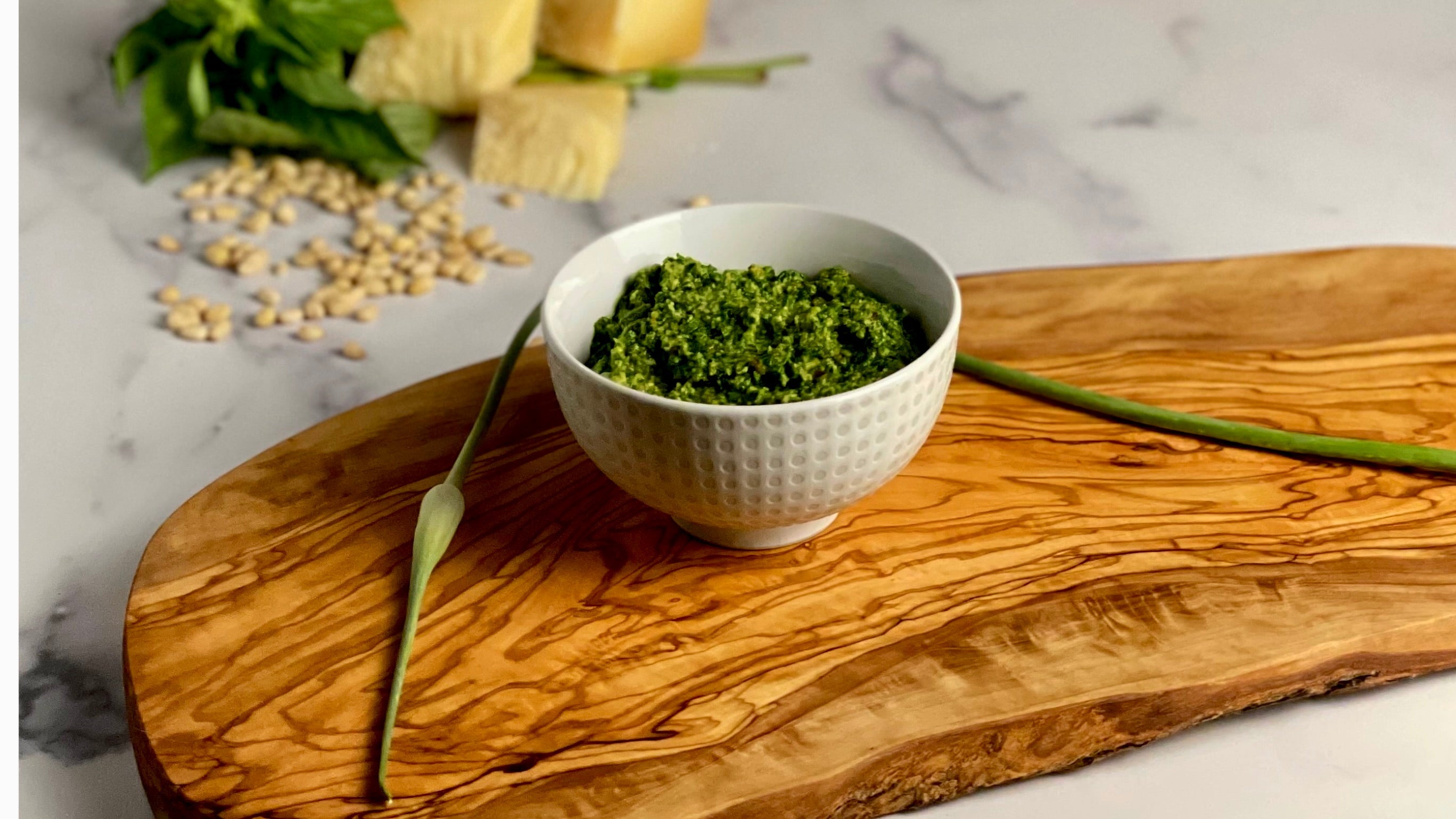 A small bowl of pesto sits on a an olive wood board in a marble kitchen. Pinenuts, basil, and parmigiano reggiano is casually placed in the background 