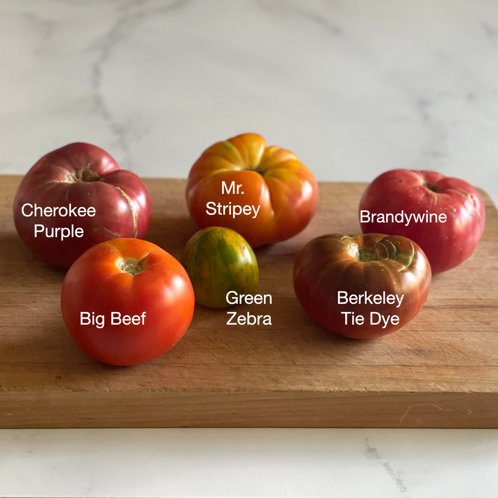 Six heirloom tomatoes are arrayed on a wooden cutting board: Cherokee Purple, Mr. Stripey, Brandywine, Big Beef, Green Zebra, Berkeley Tie Dye.