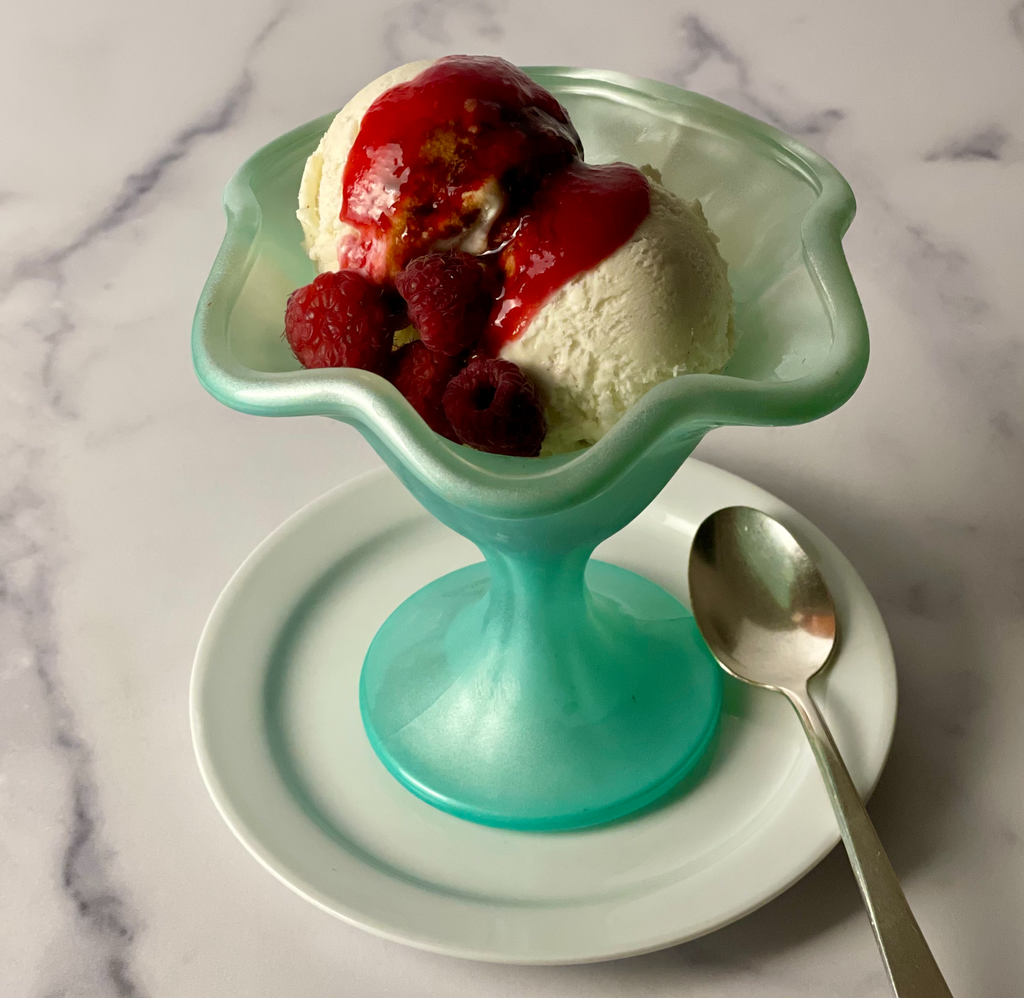 Two scoops of vanilla ice cream are visible beneath a coating of raspberry coulis, raspberry balsamic vinegar and fresh raspberries. Shown in a heavily molded vintage sundae glass with a spoon placed on a marble board.